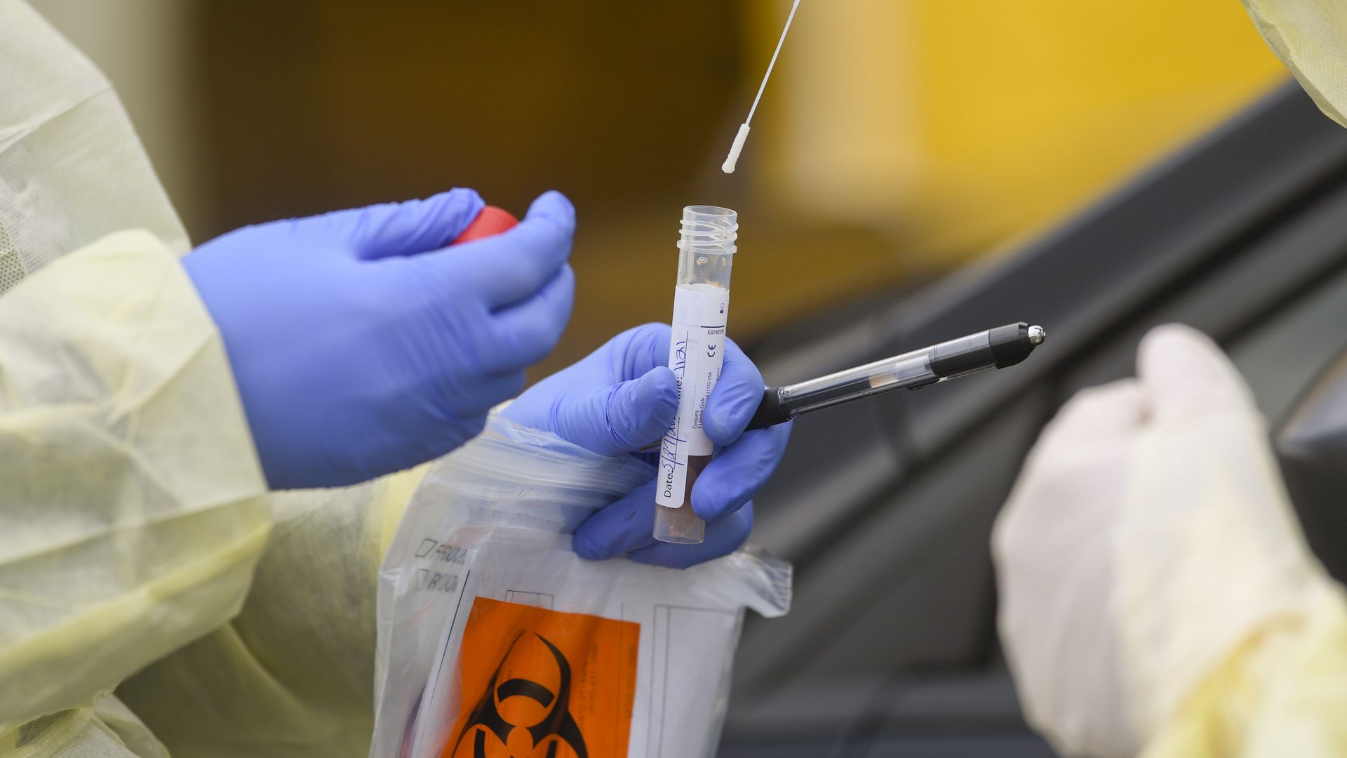 A registered nurse puts a nasal swab into a tube held by another nurse, both wearing gloves and protective gowns