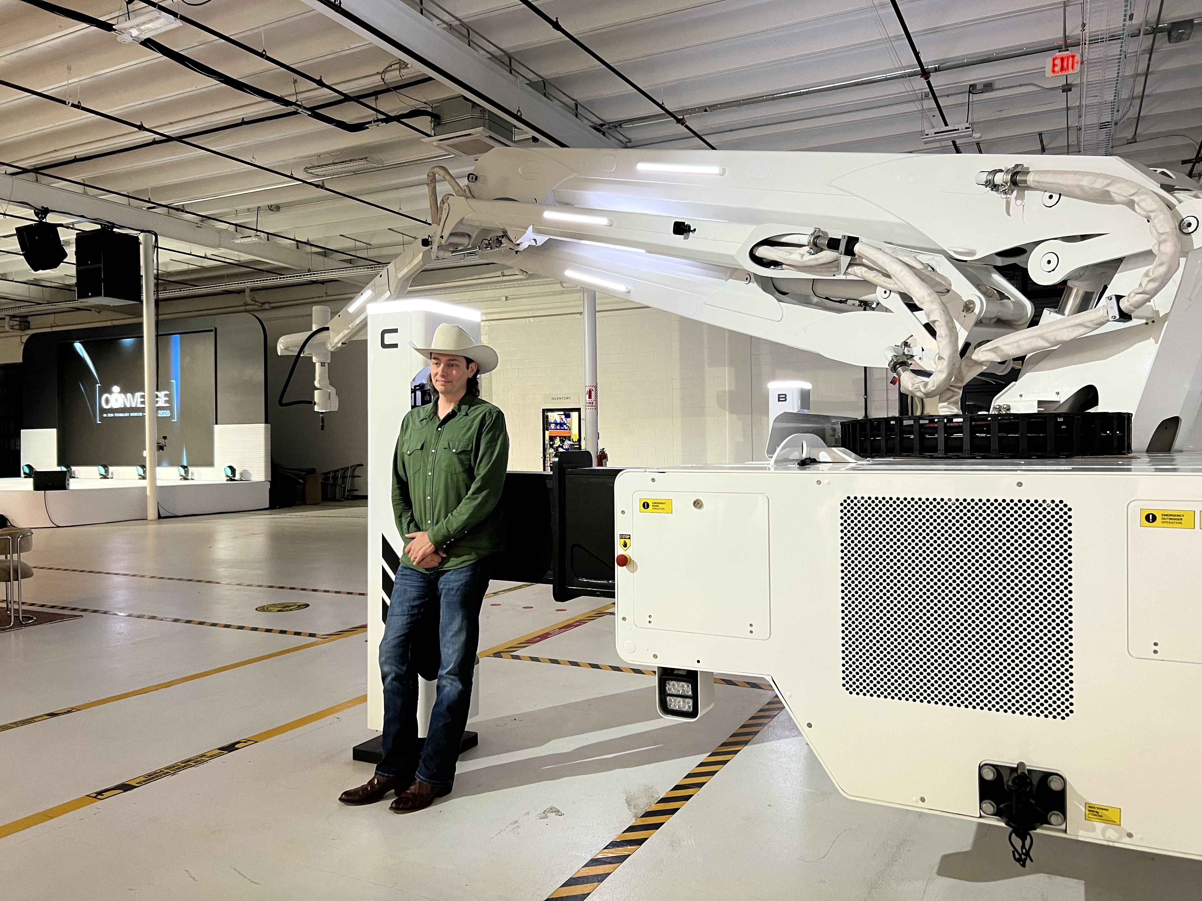 A man wearing a beige cowboy hat and green shirt stands beside a large white industrial robotic arm in a bright showroom. Exposed ceiling pipes, floor markings, and an EXIT sign are visible.