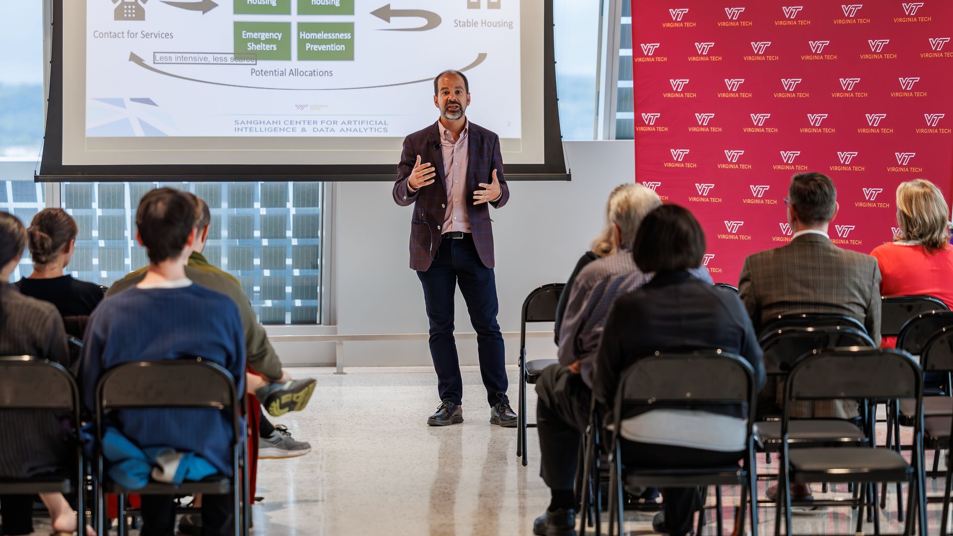 Male presenter in a plaid blazer speaks to an audience in a bright conference room; a projection screen shows a chart on the left, while a Virginia Tech burgundy backdrop lines the right as attendees listen.