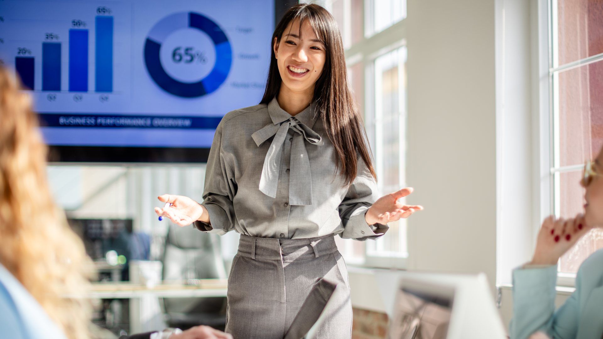 A smiling businesswoman wearing a gray blouse with a bow and gray skirt presents in a bright office. A large screen shows charts and a 65% donut graph while colleagues listen.