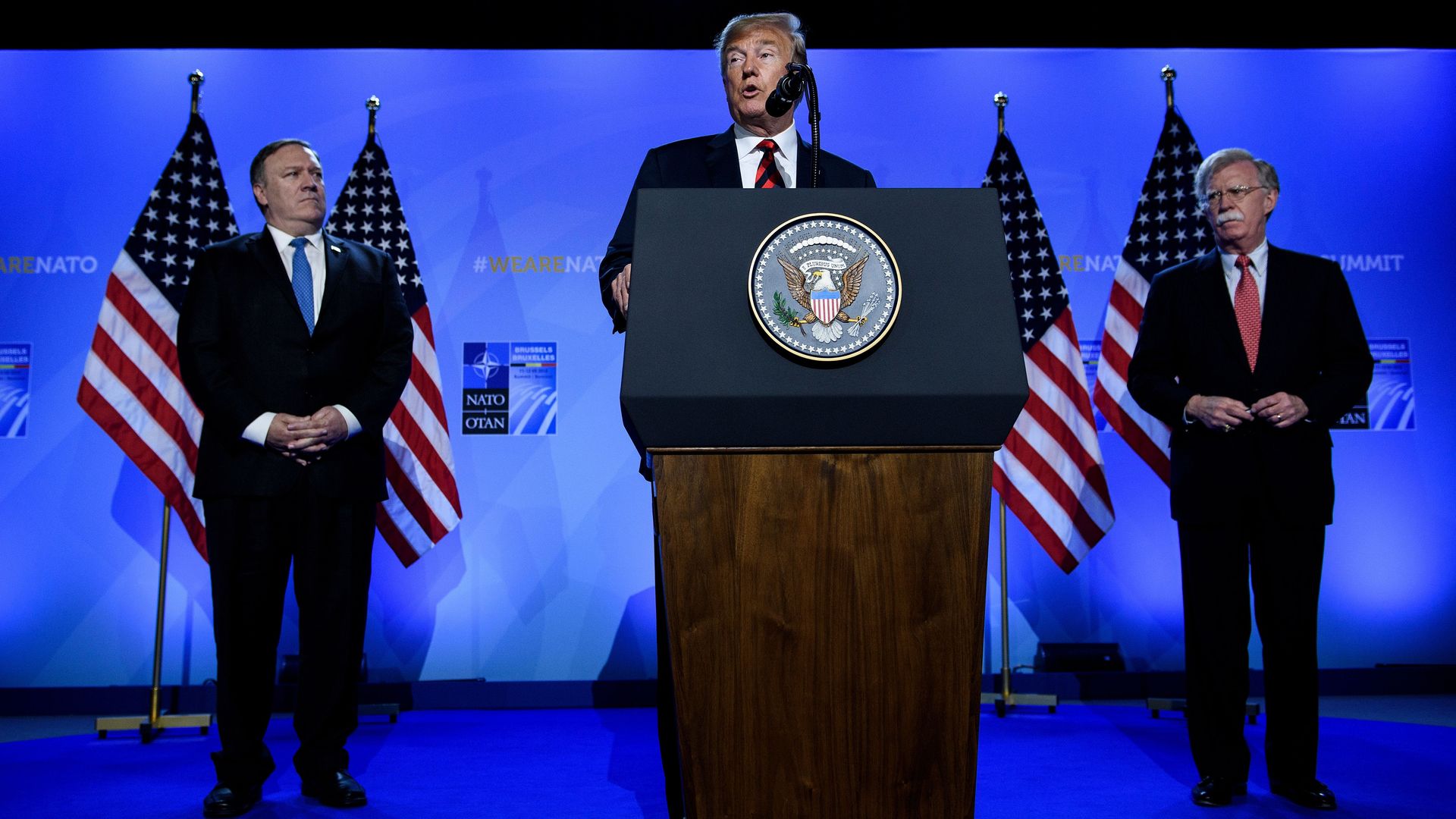 Mike Pompeo and John Bolton on stage behind Donald Trump, speaking at a lectern