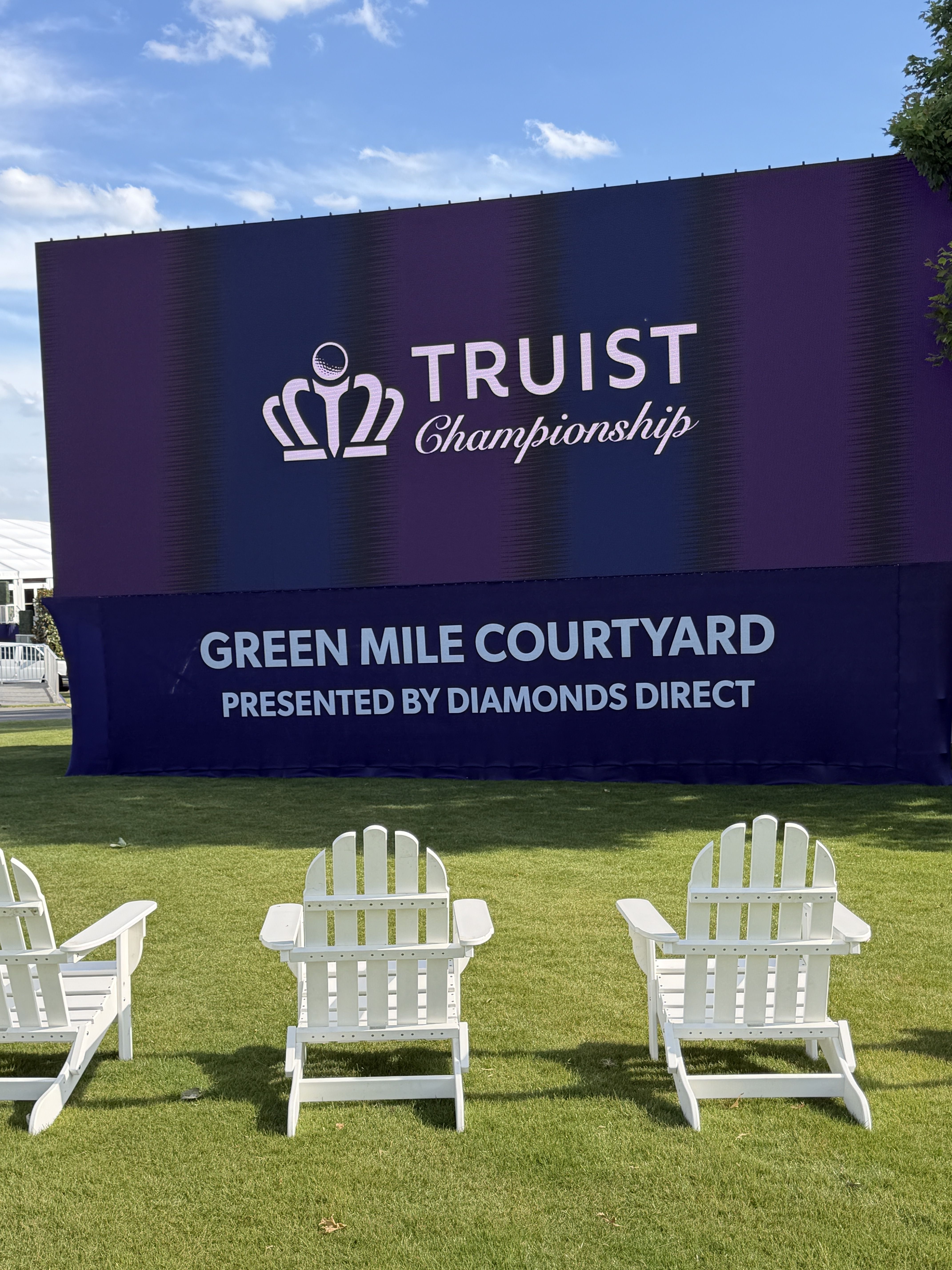 Outdoor scene with a large purple banner reading TRUIST Championship; crown logo, white Adirondack chairs on green grass, blue sky with a few clouds, and a tree on the right.