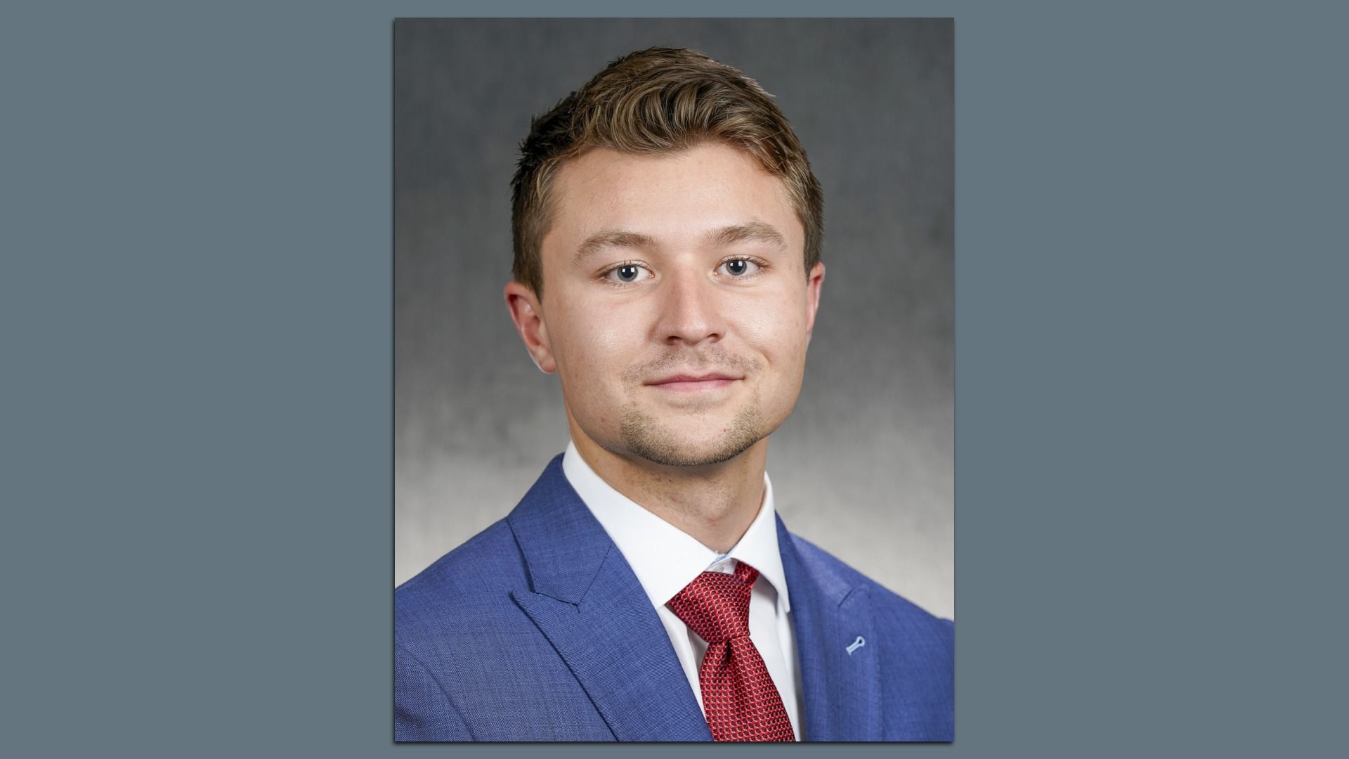 Portrait of a young man with short light brown hair wearing a blue suit, white shirt, and red tie, looking at the camera with a slight smile, against a gray gradient background.