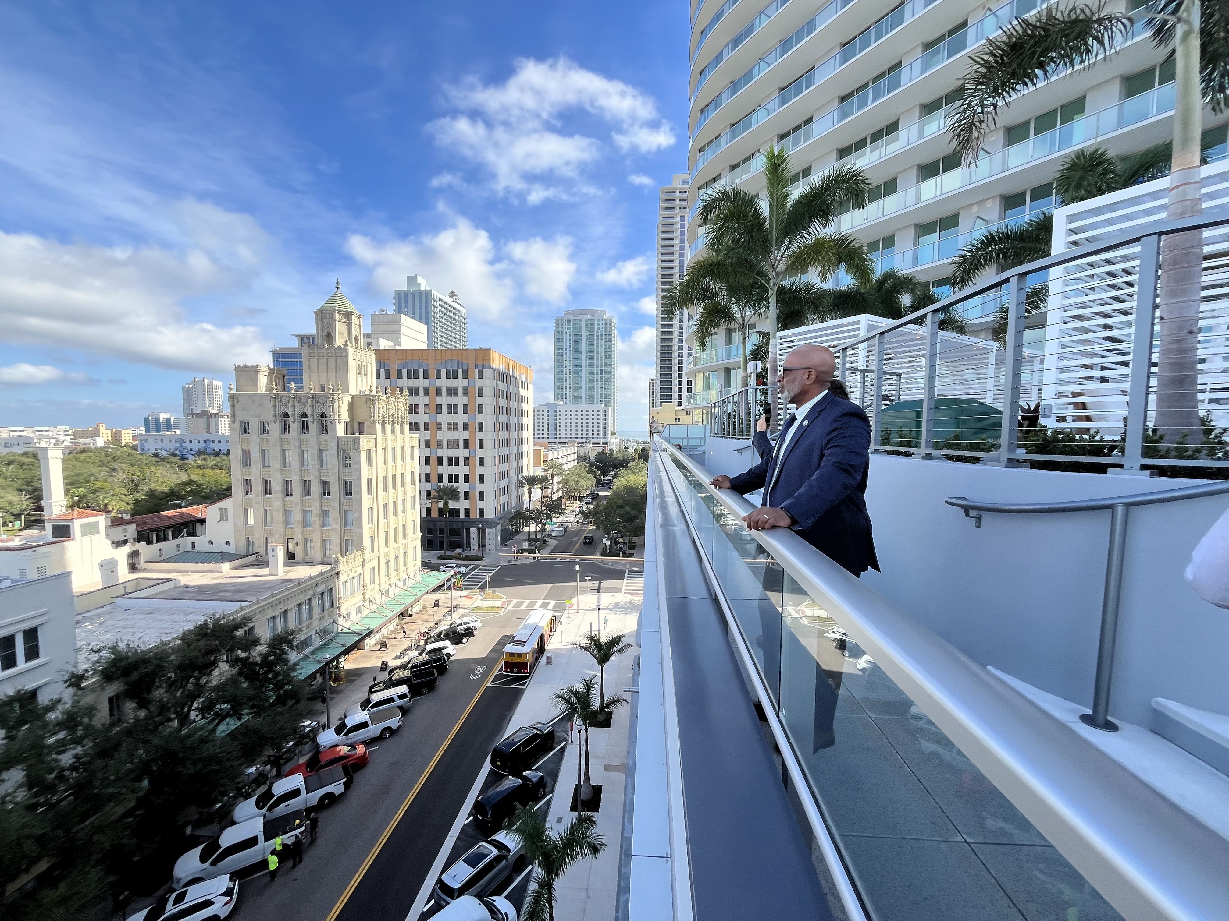 Man in a blue suit stands on a balcony overlooking a sunny city street with parked cars and tall buildings under a blue sky with clouds.