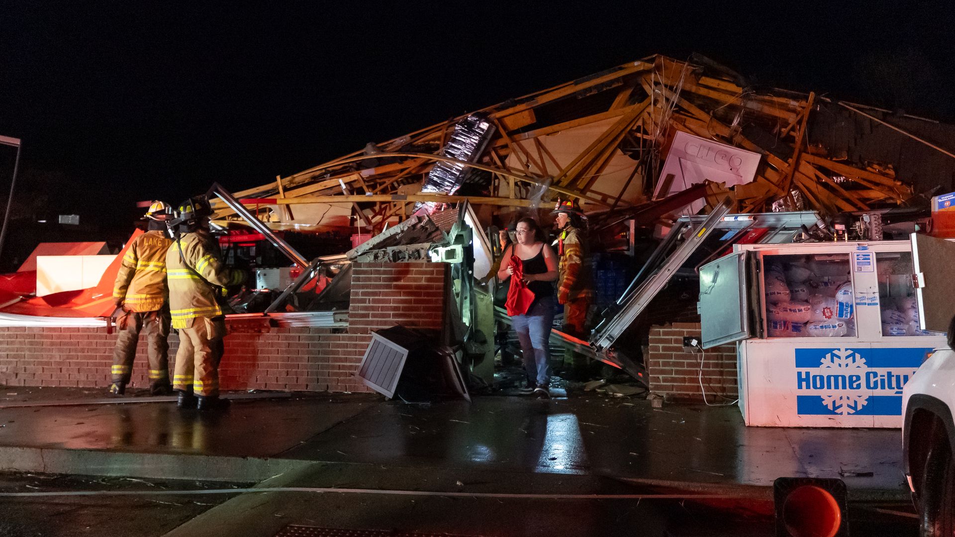 Night scene of a collapsed storefront with firefighters in gear, debris and wooden beams; a woman in jeans holds a red cloth amid damaged equipment and a Home City freezer.