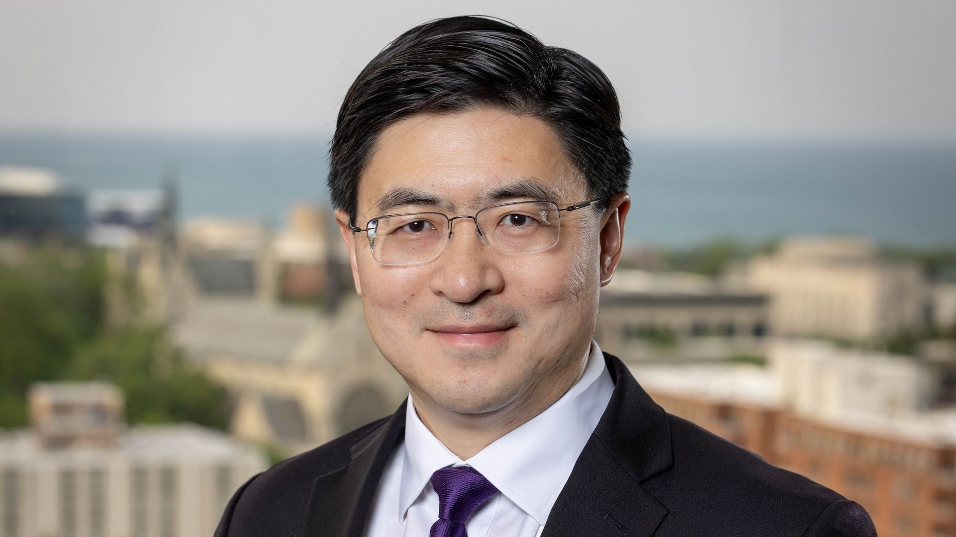 Close-up portrait of a man in a dark suit and purple tie, wearing glasses, outdoors with a blurred city and water background.