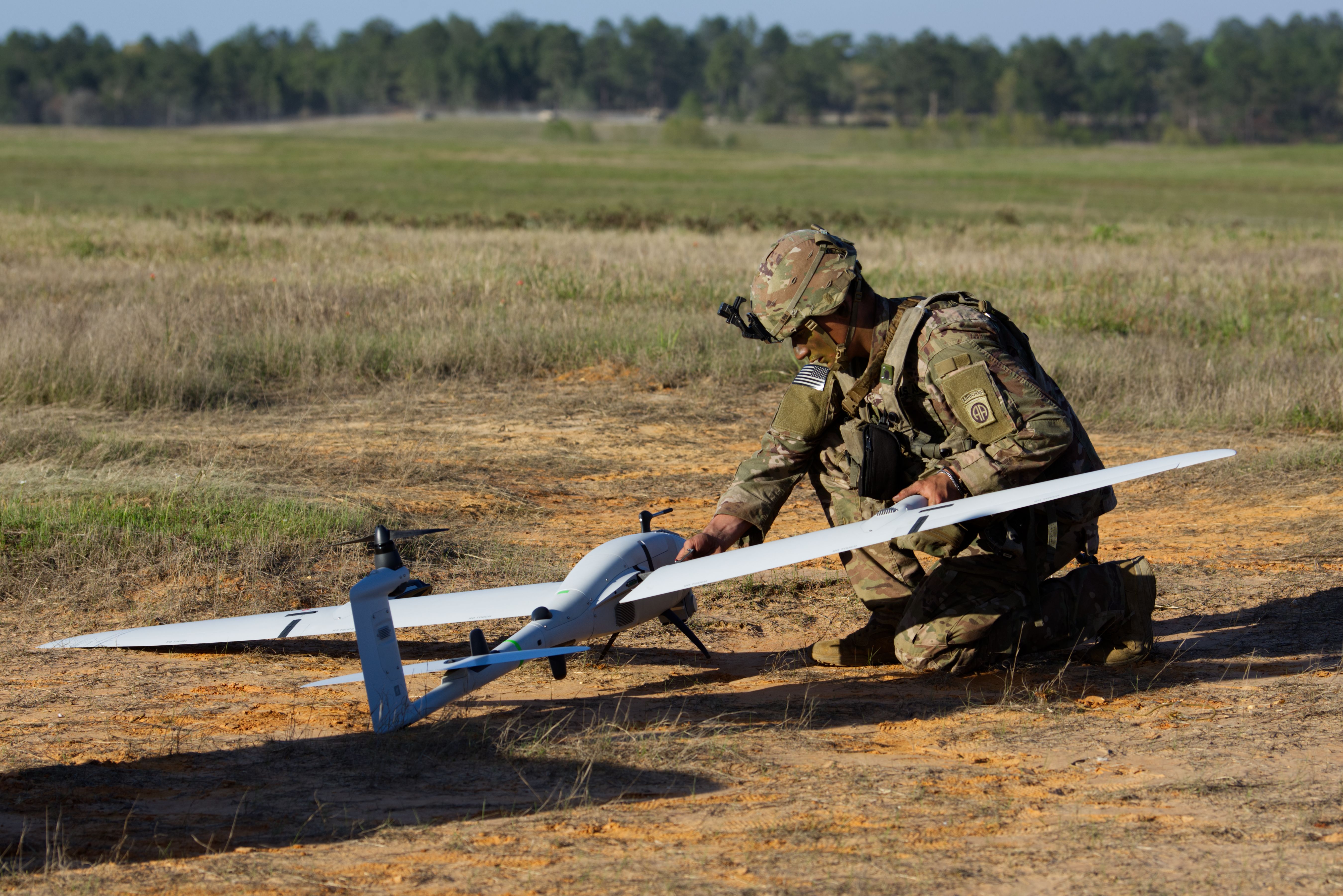Camouflage-suited soldier kneels in a dry field, inspecting a white fixed-wing drone with long wings. He wears a helmet and tactical gear, with an American flag patch visible on his sleeve.