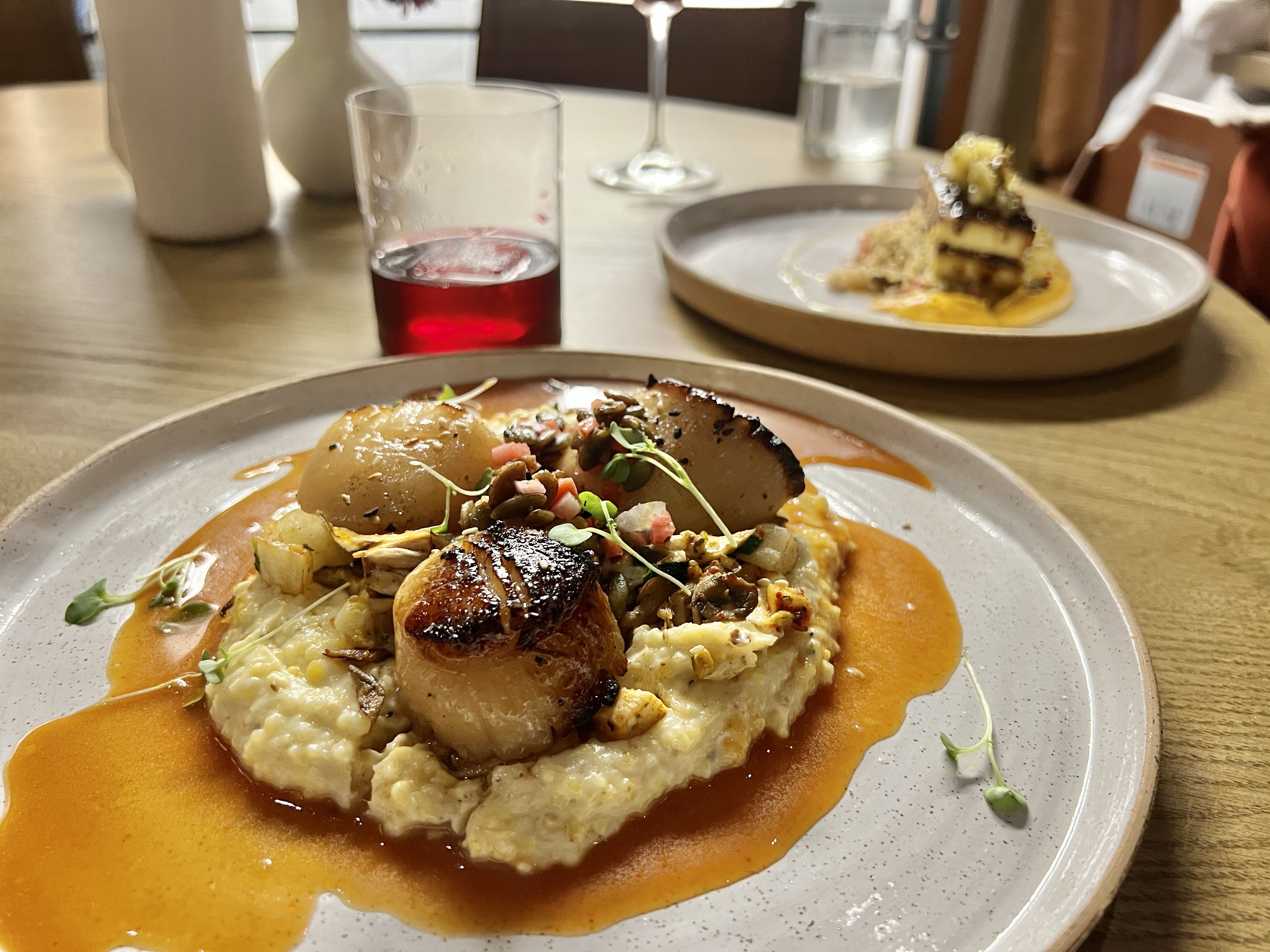 Seared scallop over risotto with microgreens and seeds, orange sauce around; back plates show a dessert slice and drinks on a wooden table.