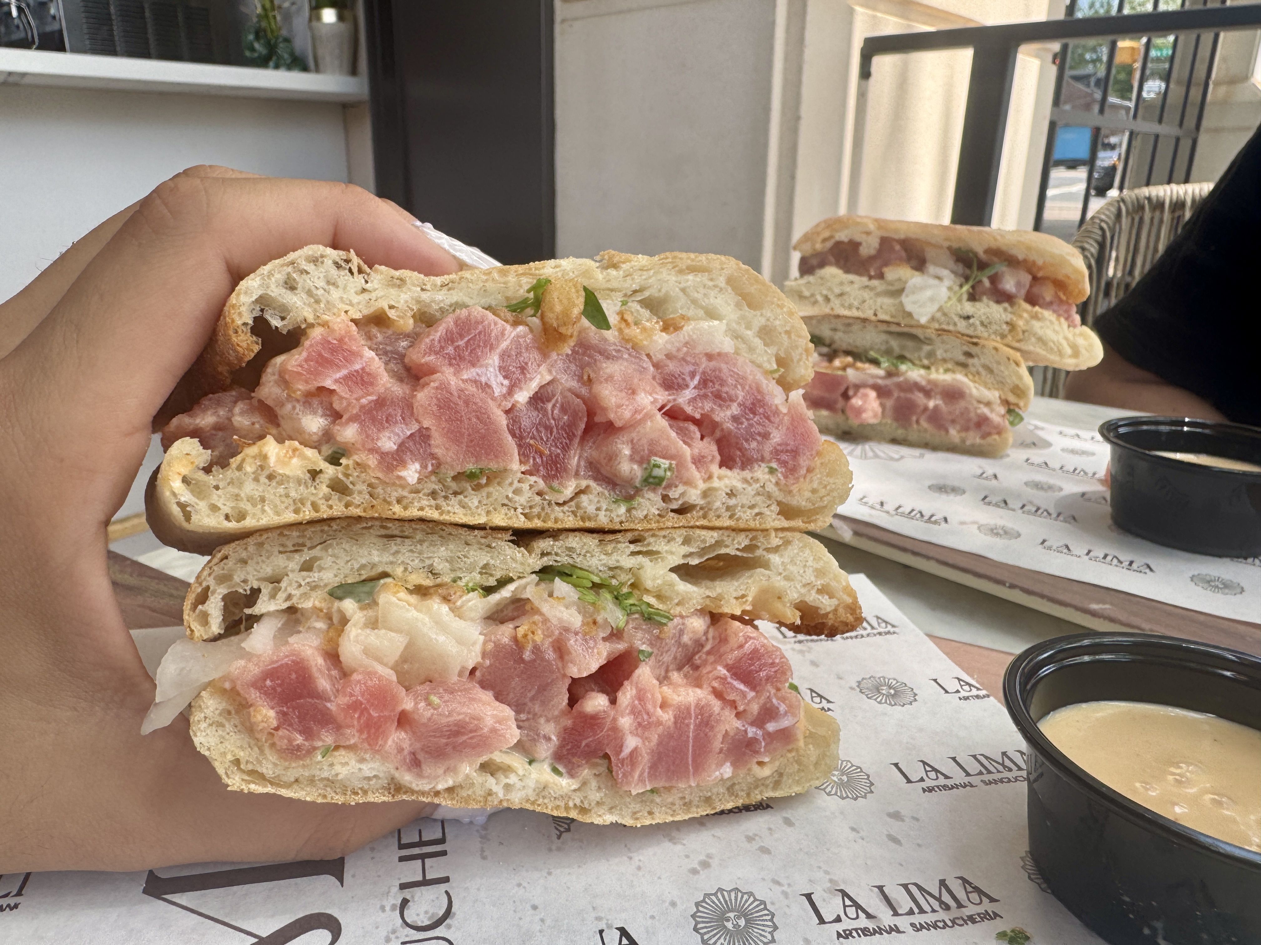 Outdoors, a hand holds a crusty sandwich with chunky pink meat, herbs, and soft bread; another half sits on the table with dipping cups and a railing in the background.