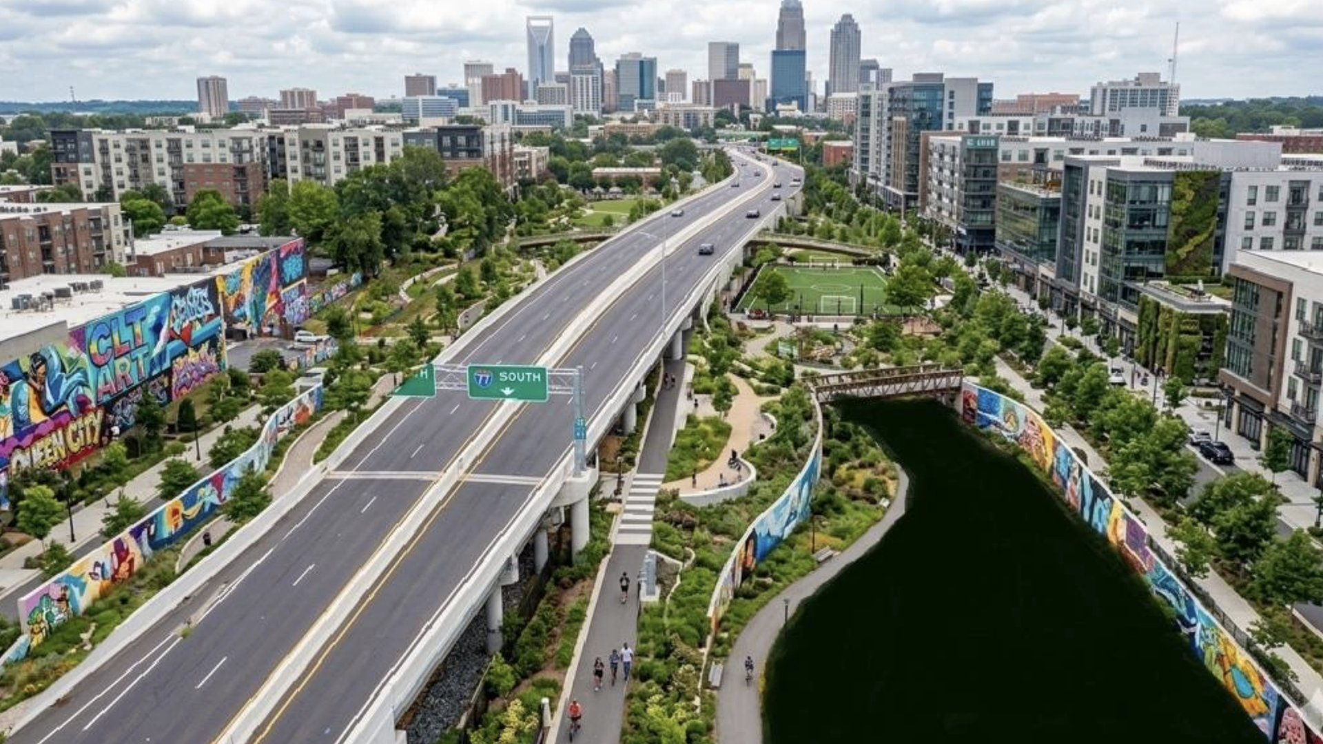 An aerial city view with a raised highway curving over a mural-lined wall and green park along a canal; pedestrians and cyclists on paths; distant skyline under a cloudy sky.
