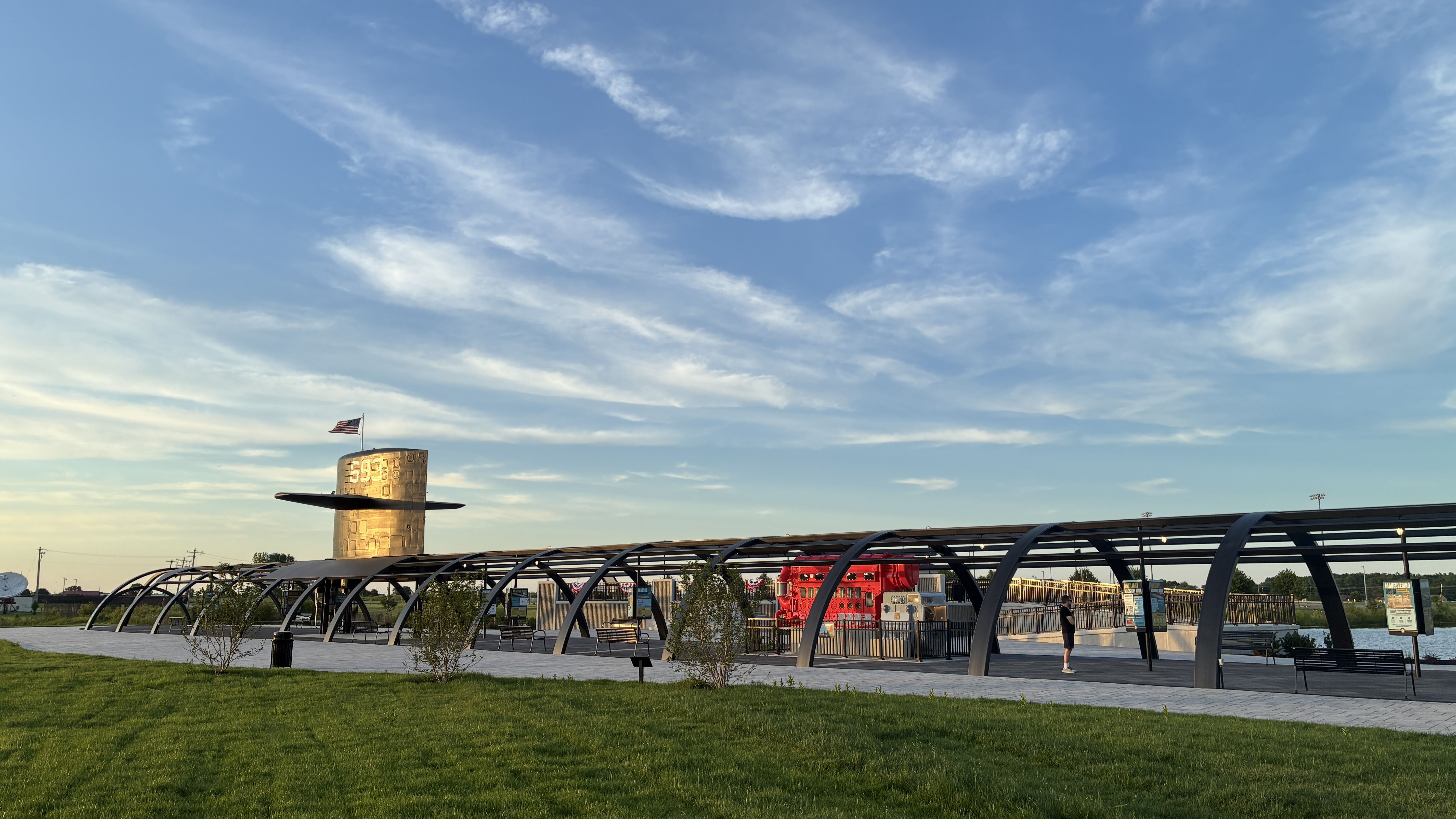 Wide view of a sunny park with a curved metal walkway and a cylindrical tower topped by an American flag; green grass in the foreground and wispy clouds in a blue sky.