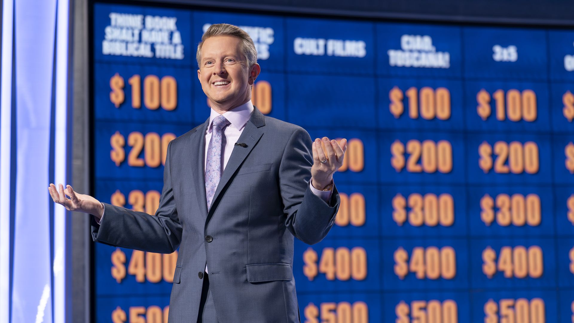 Ken Jennings in a dark gray suit stands with his hands gesturing to his sides in front of the Jeopardy! board with dollar numbers on it.