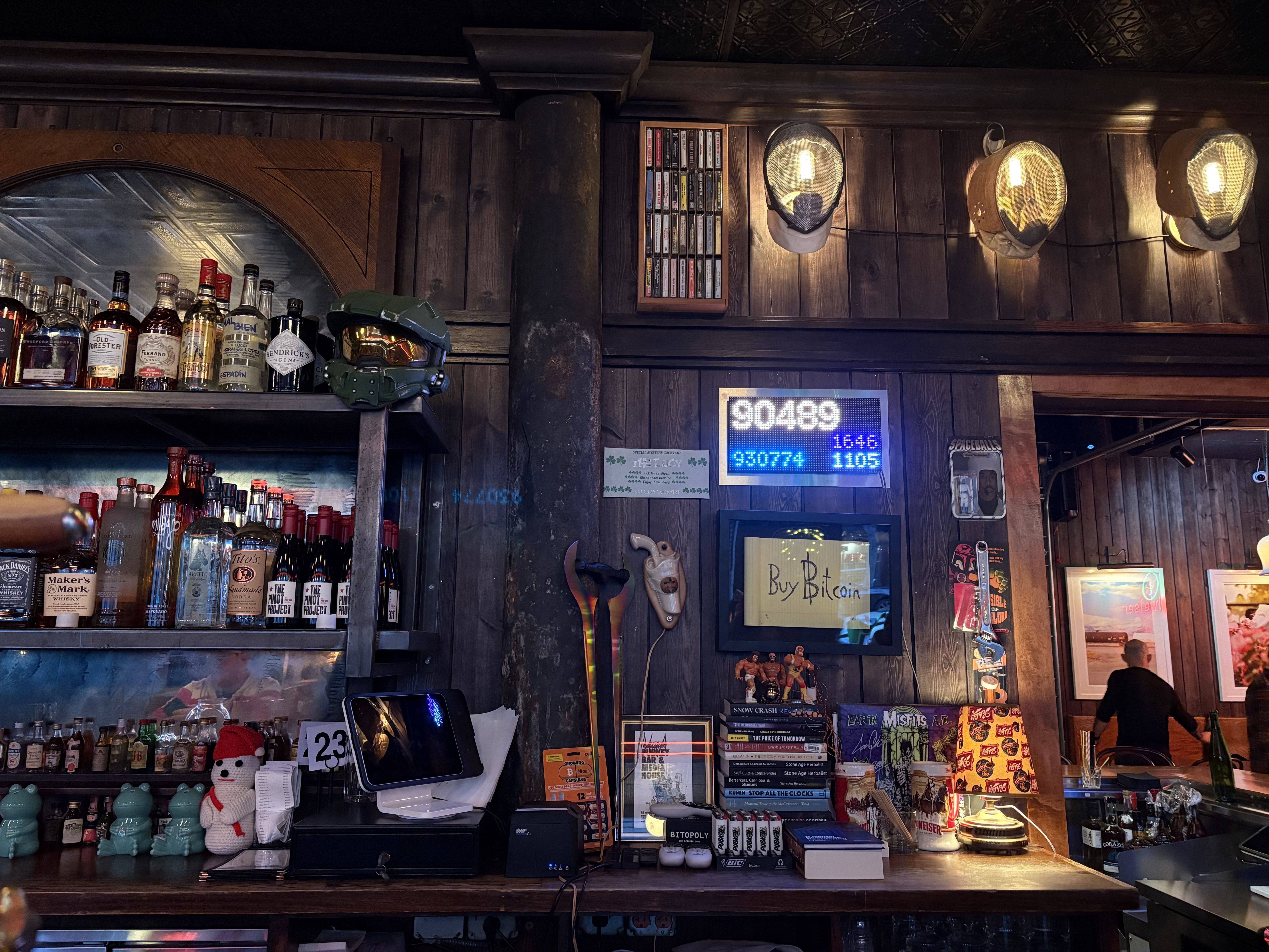 Bar interior with wooden walls, bottles of liquor, a green helmet on a shelf, figurines, books, and a digital Bitcoin price display above a sign reading "Buy Bitcoin." Warm lighting.