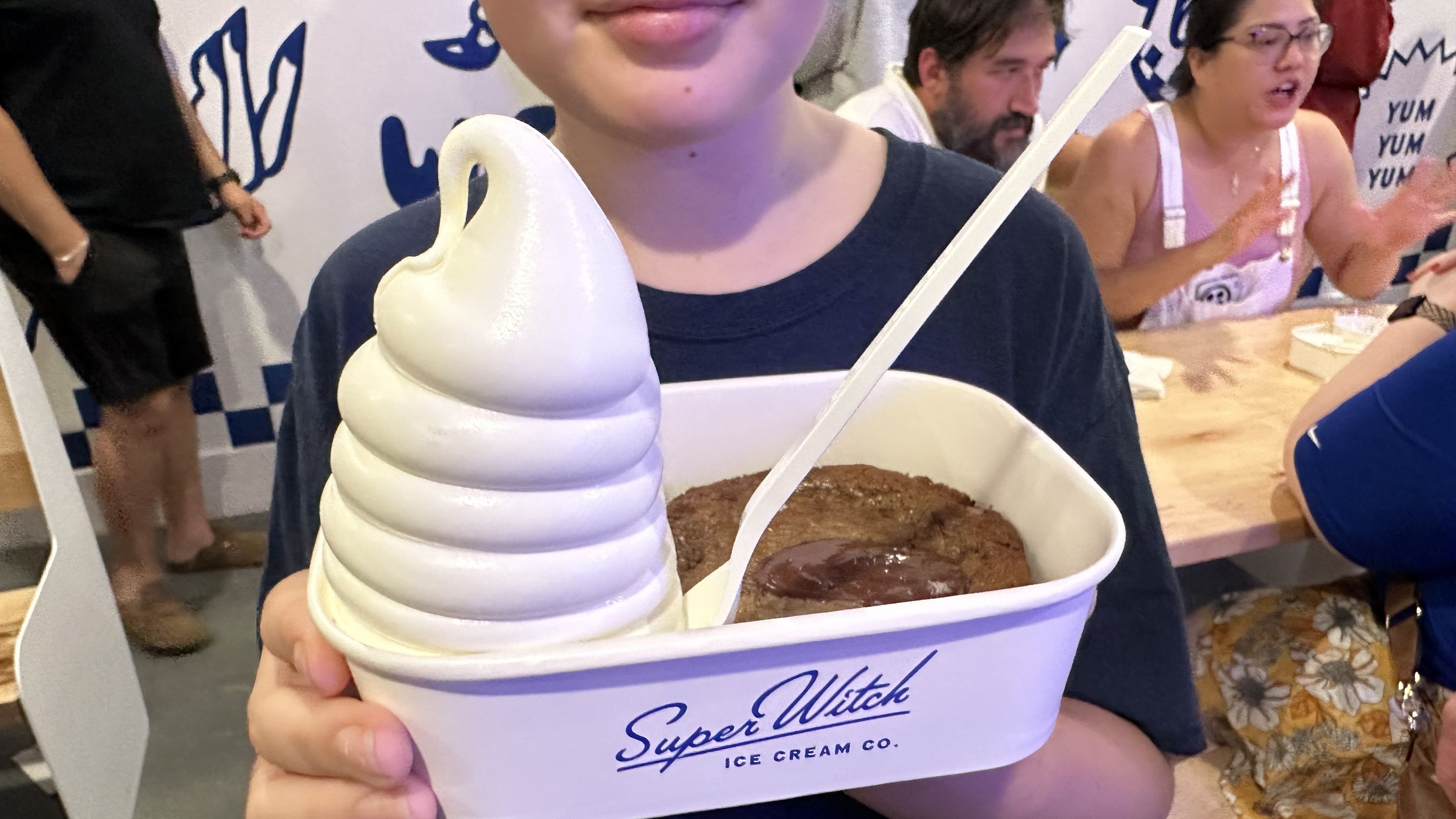A person in a dark shirt holds a cup topped with a tall white soft-serve ice cream swirl; chocolate ice cream is visible in the cup, with others seated at a counter in the background.