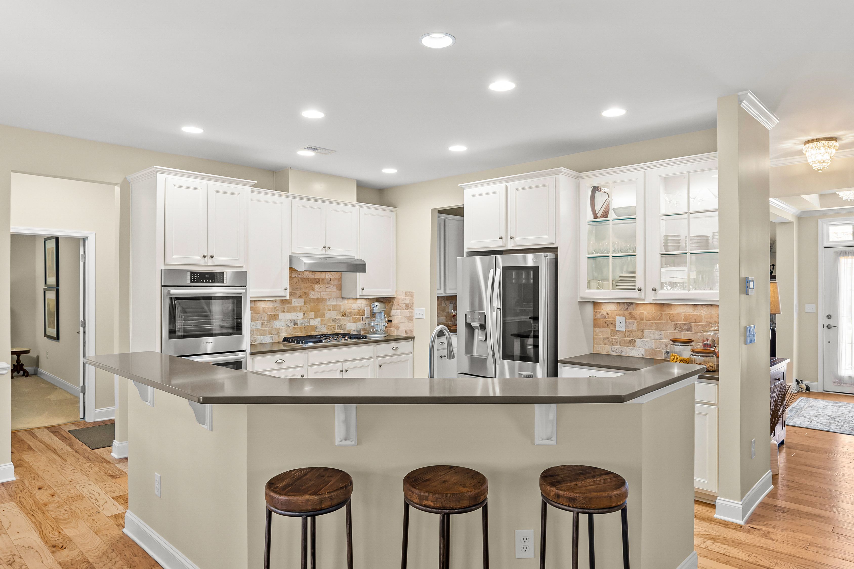 Bright, open kitchen with white cabinets, beige walls, and a large central island. Three wooden stools at the counter, stainless steel appliances, glass-front cabinets, and a tan brick backsplash.