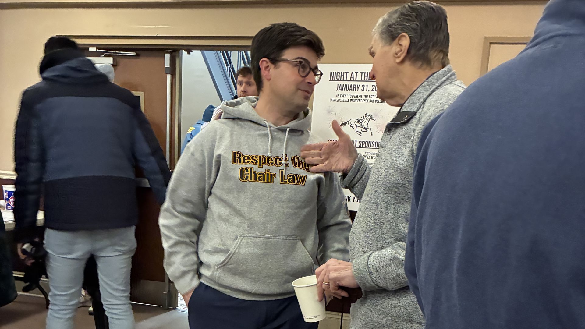 Two men engaged in conversation indoors; one wearing glasses and a grey hoodie stating "Respect the Chair Law," the other gesturing and holding a white paper cup, with a poster in the background.