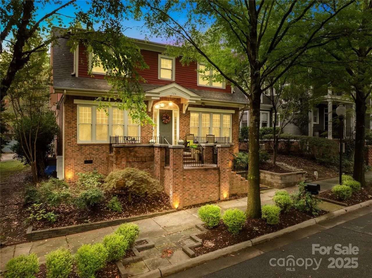 Two-story brick and red siding house with lit windows and front porch, surrounded by green trees and bushes along a sidewalk, taken at dusk. "For Sale" sign in corner.