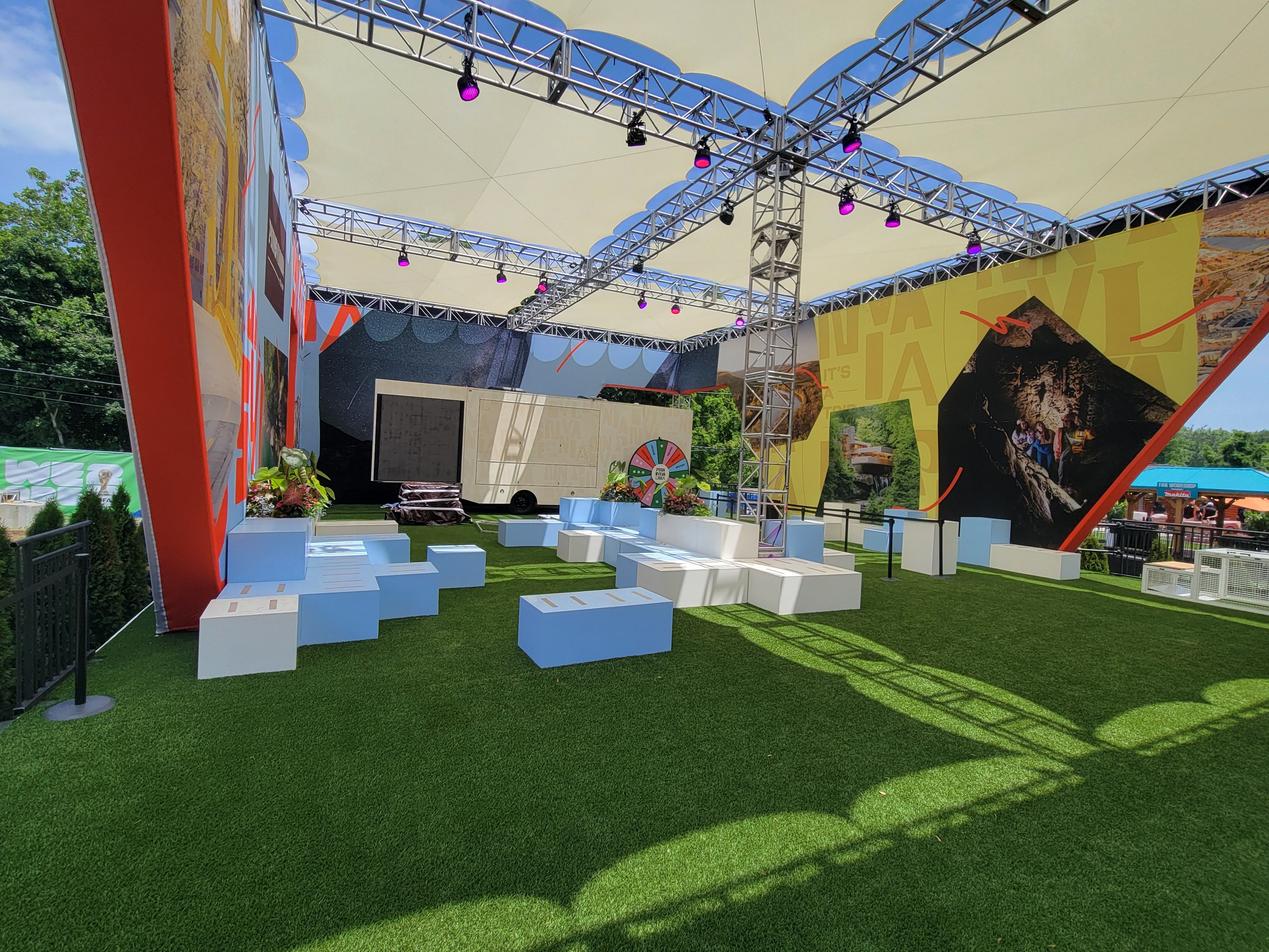 Outdoor stage with a beige canvas roof, a metal truss lighting rig with purple stage lights, colorful wall panels depicting mountains; artificial grass, white benches, and potted plants.
