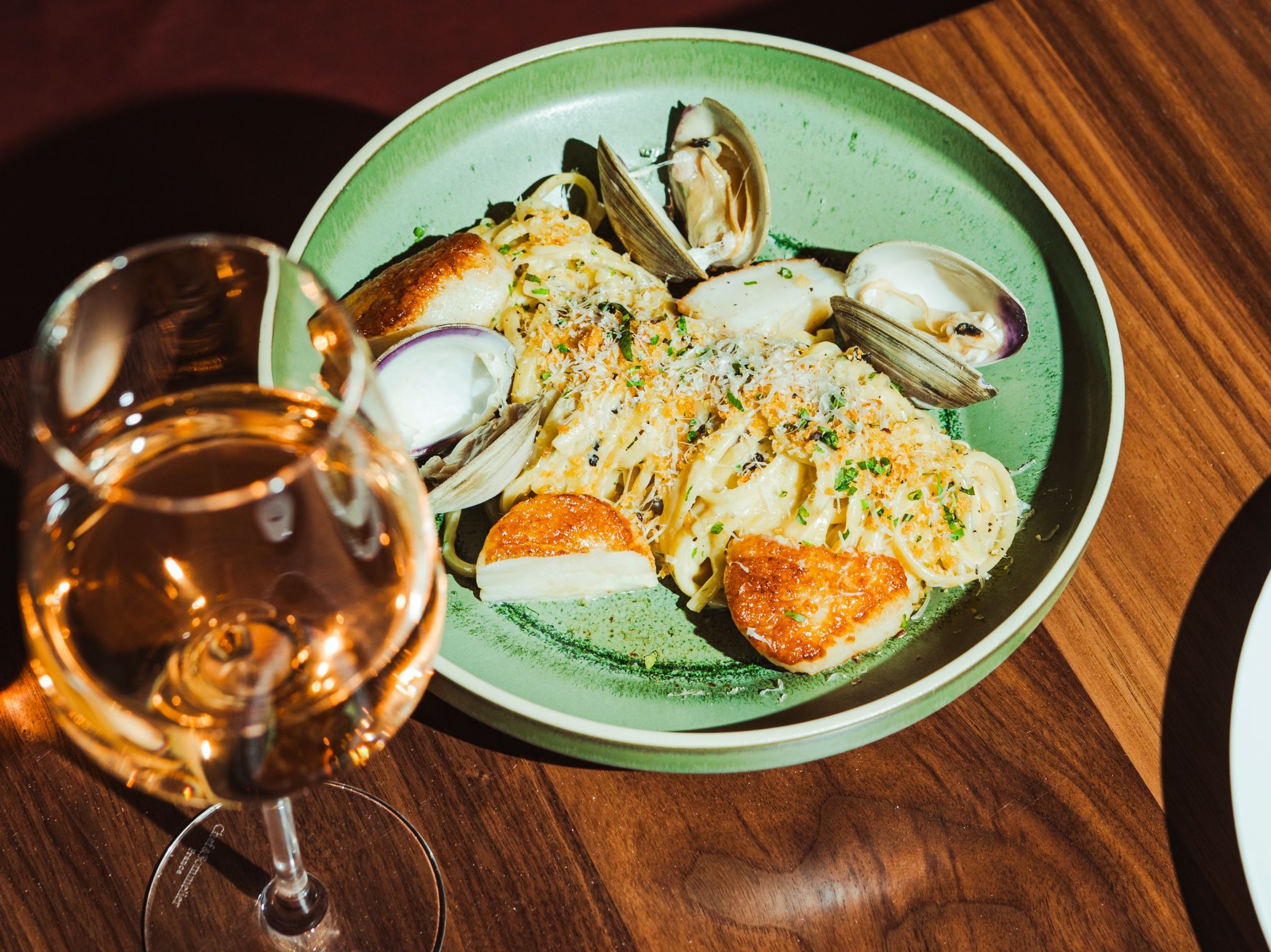Top-down view of a green plate of seafood pasta with clams, breadcrumbs, and herbs on a wooden table; a wine glass is in the foreground.