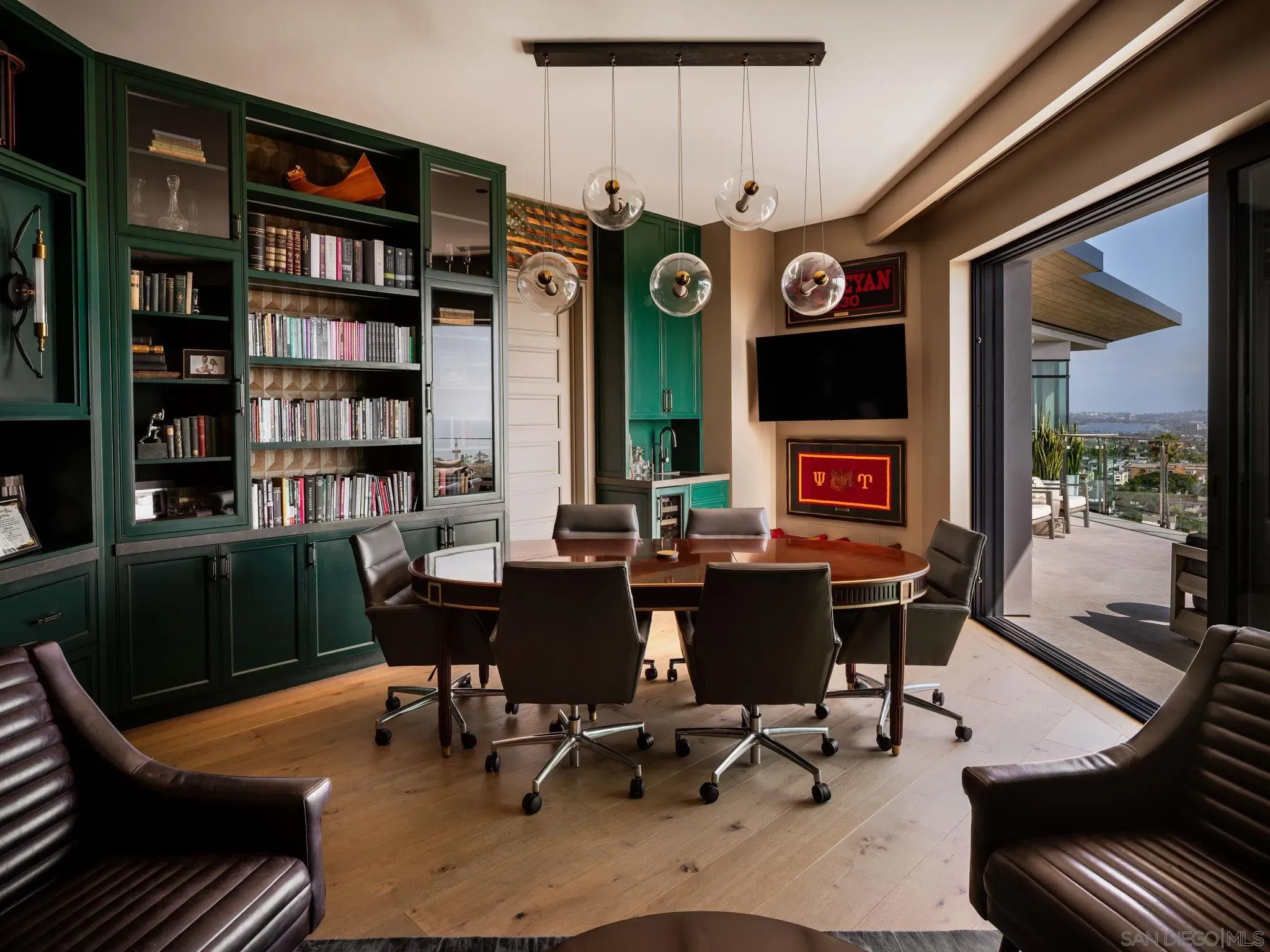Luxurious home office with dark green built-in bookshelves, glass-front cabinets, a round wooden table and six swivel chairs, five glass globe pendant lights, and a balcony view of the city.
