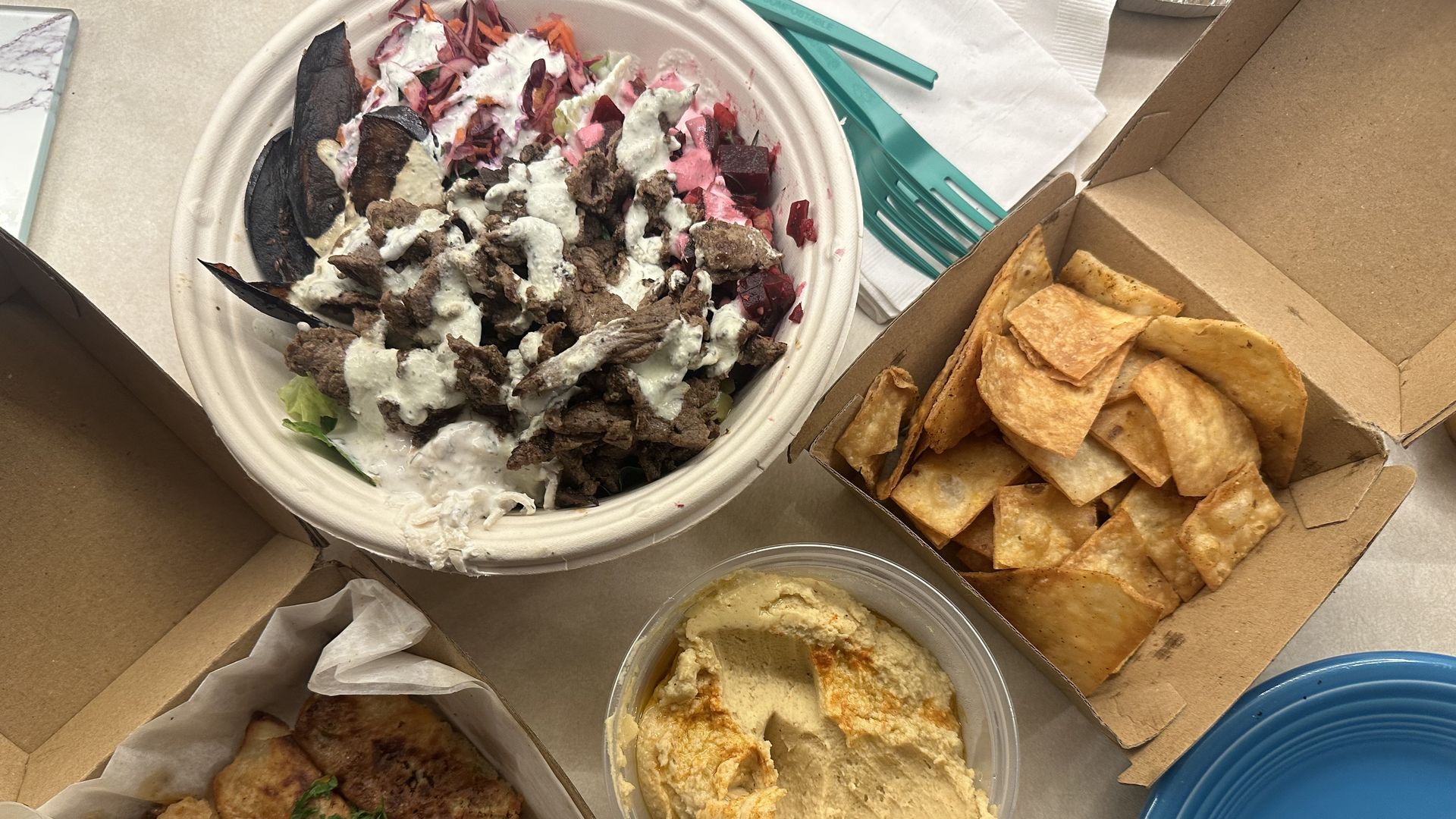 Top-down view of a takeout meal: a bowl of beef with creamy sauce and veggie toppings, beet bits, a box of tortilla chips, hummus cup, and teal forks near a blue plate.