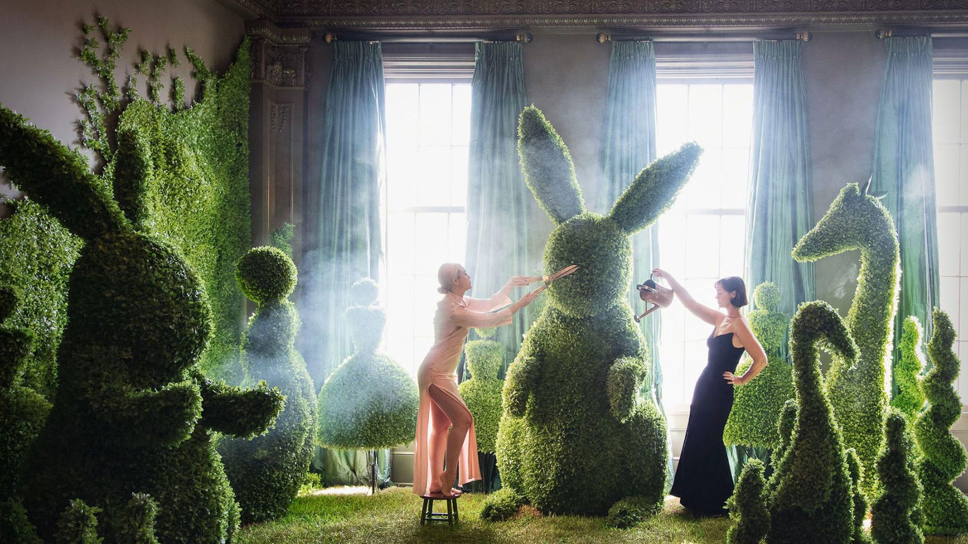 Two women in elegant dresses care for large green topiary sculptures shaped like animals and figures in a grand room with ornate ceiling and tall windows with green curtains.