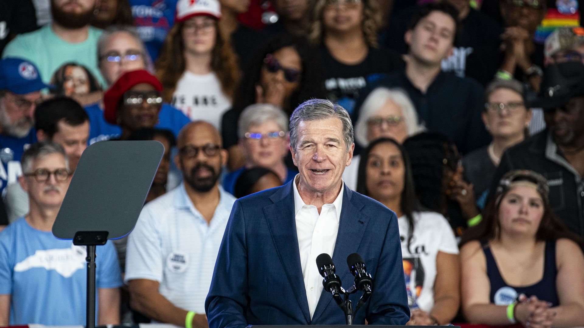 Man in a blue suit and white shirt speaking at a podium with microphone, surrounded by a diverse crowd of people behind him, some wearing glasses and hats, indoors.