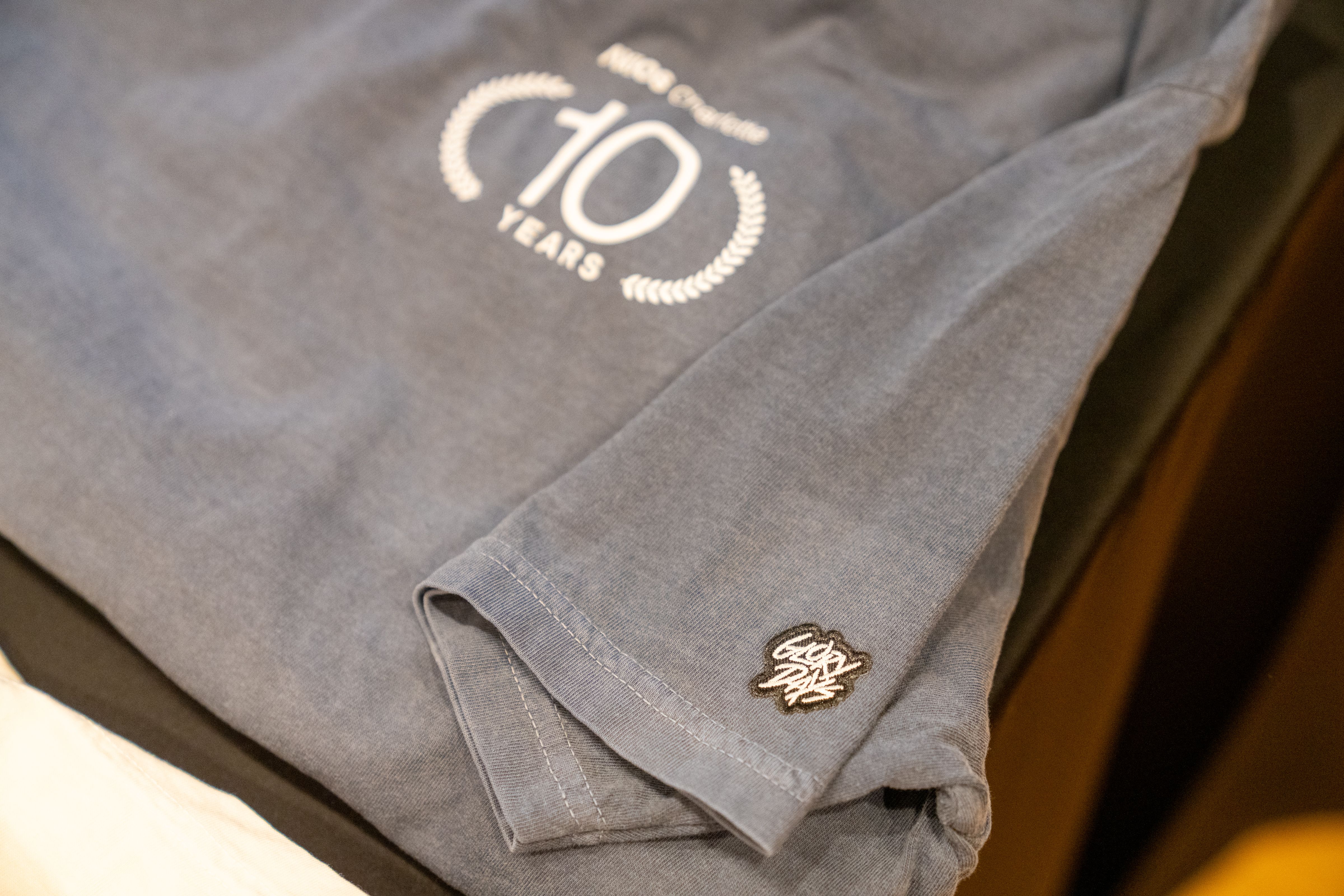 Close-up of a gray shirt with a "10 Years" anniversary logo in white and a small embroidered black and white patch on the sleeve cuff.