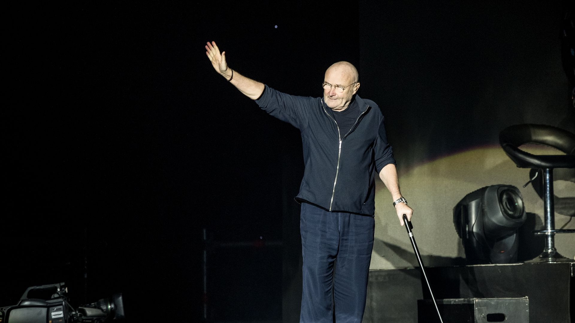 An older, bald man in a navy jacket and glasses stands on a dark stage, raising his right hand while holding a microphone in his left; stage lights and equipment are visible nearby.