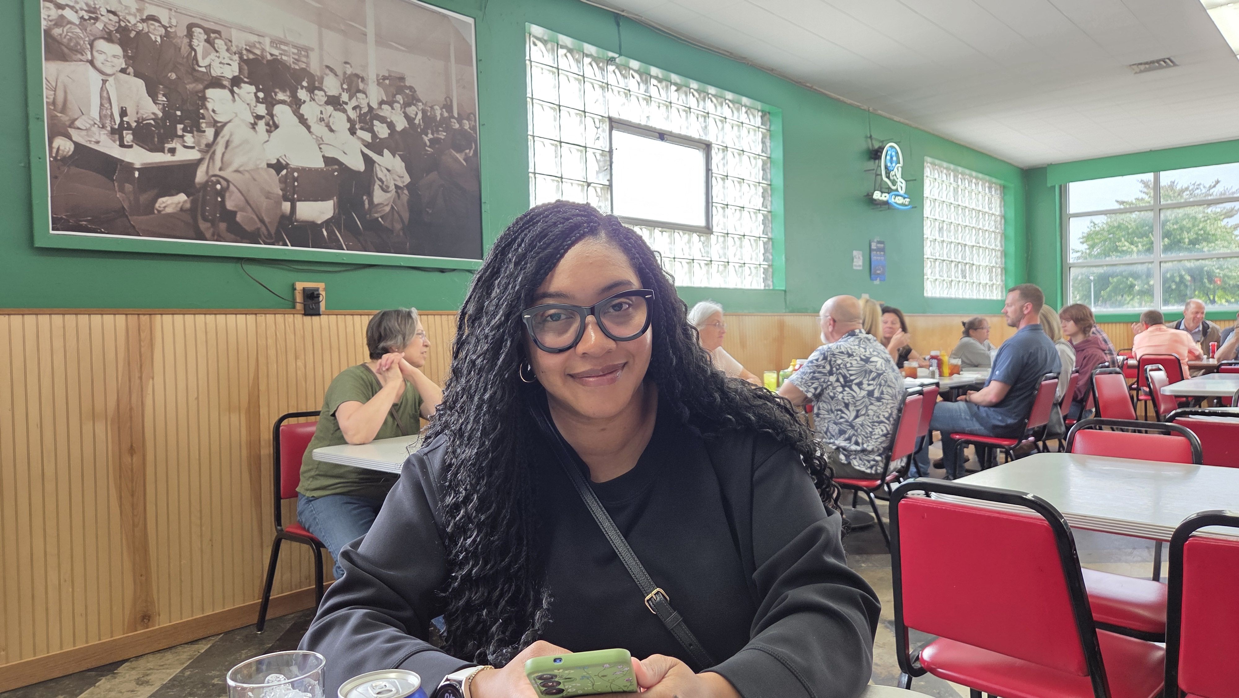 A smiling woman with glasses sits at a table in a bright diner with green walls and red chairs. A large black-and-white photo hangs on the wall behind her, and diners chat at other tables.