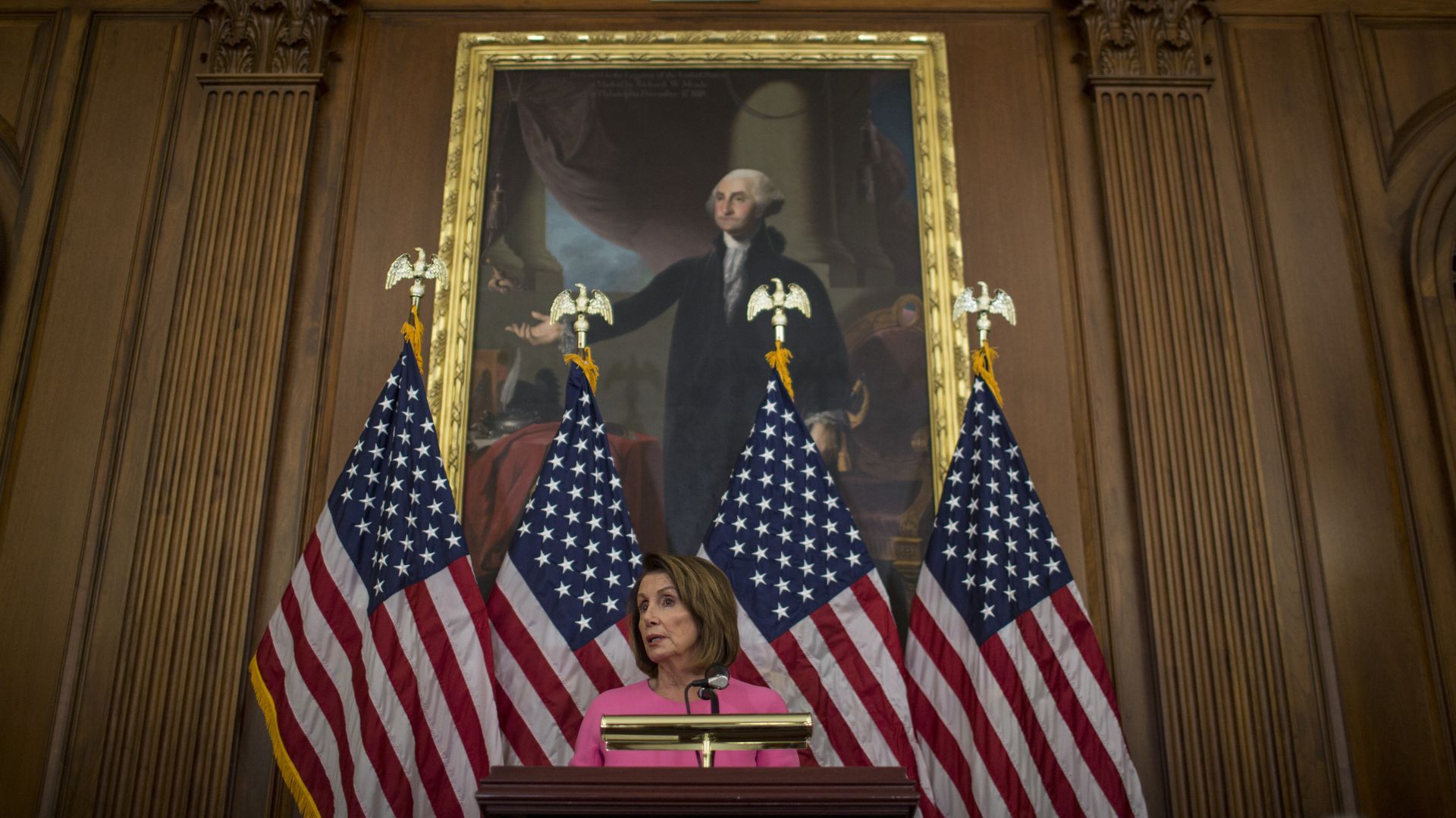 Nancy Pelosi speaks from a podium in front of American flags and a painting of George Washington