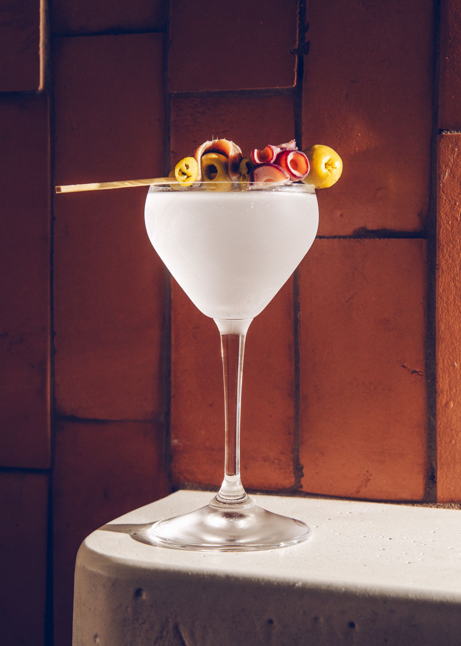 Clear cocktail in a stemmed glass garnished with olives and red octopus tentacles, set against a reddish-brown brick wall on a light-colored surface.
