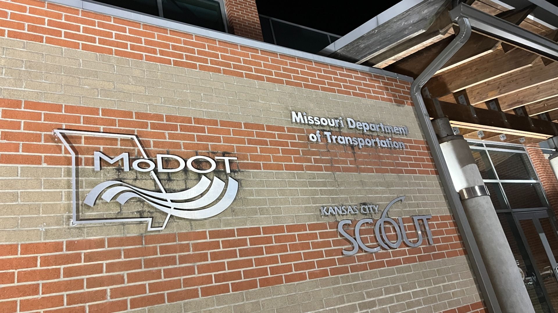Brick wall with silver signage for Missouri Department of Transportation, MoDOT logo featuring the state outline and waves, and Kansas City Scout logo near an entrance with wooden beams and columns.