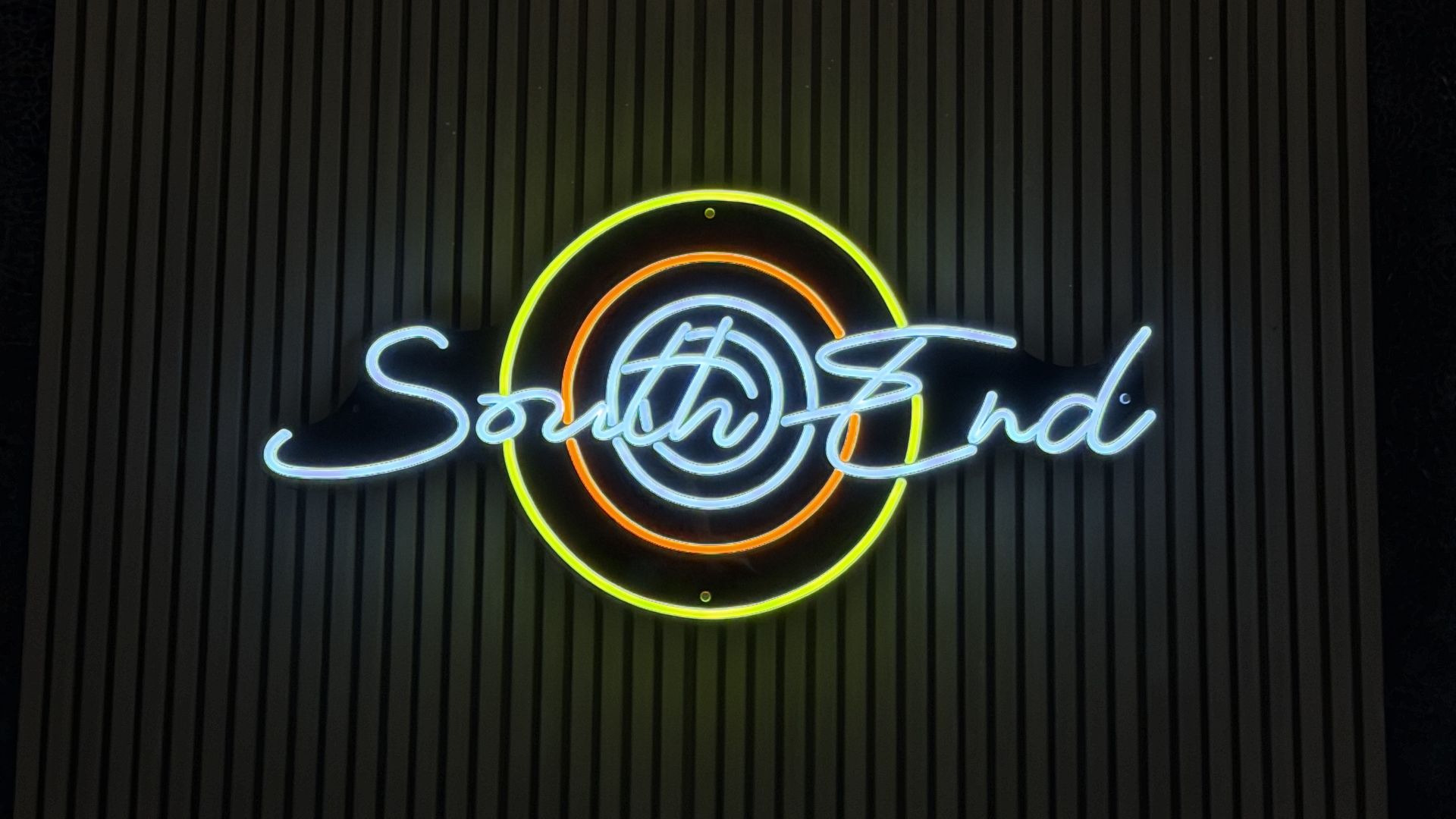 Dim interior featuring large woven wicker globes with warm light, dark striped wall, and a neon sign reading "Southend" with concentric circles in neon green, yellow, orange, and blue.
