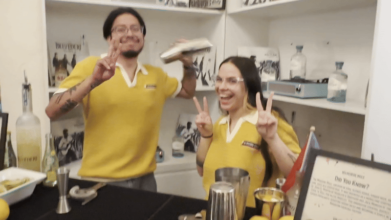 Two bartenders in yellow shirts behind a bar counter, smiling and making peace signs with hands, surrounded by bottles and cocktail tools in a white room.