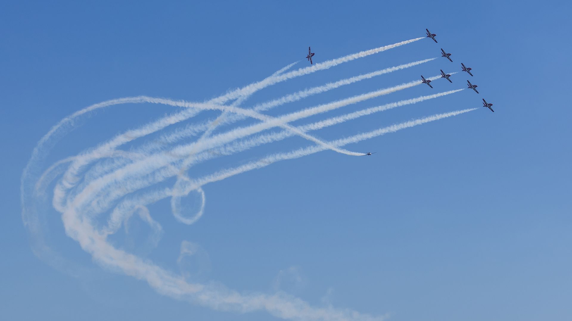 Nine planes in formation creating looping smoke trails above a long pier with a red-roofed building, blue ocean waves, and clear blue sky.