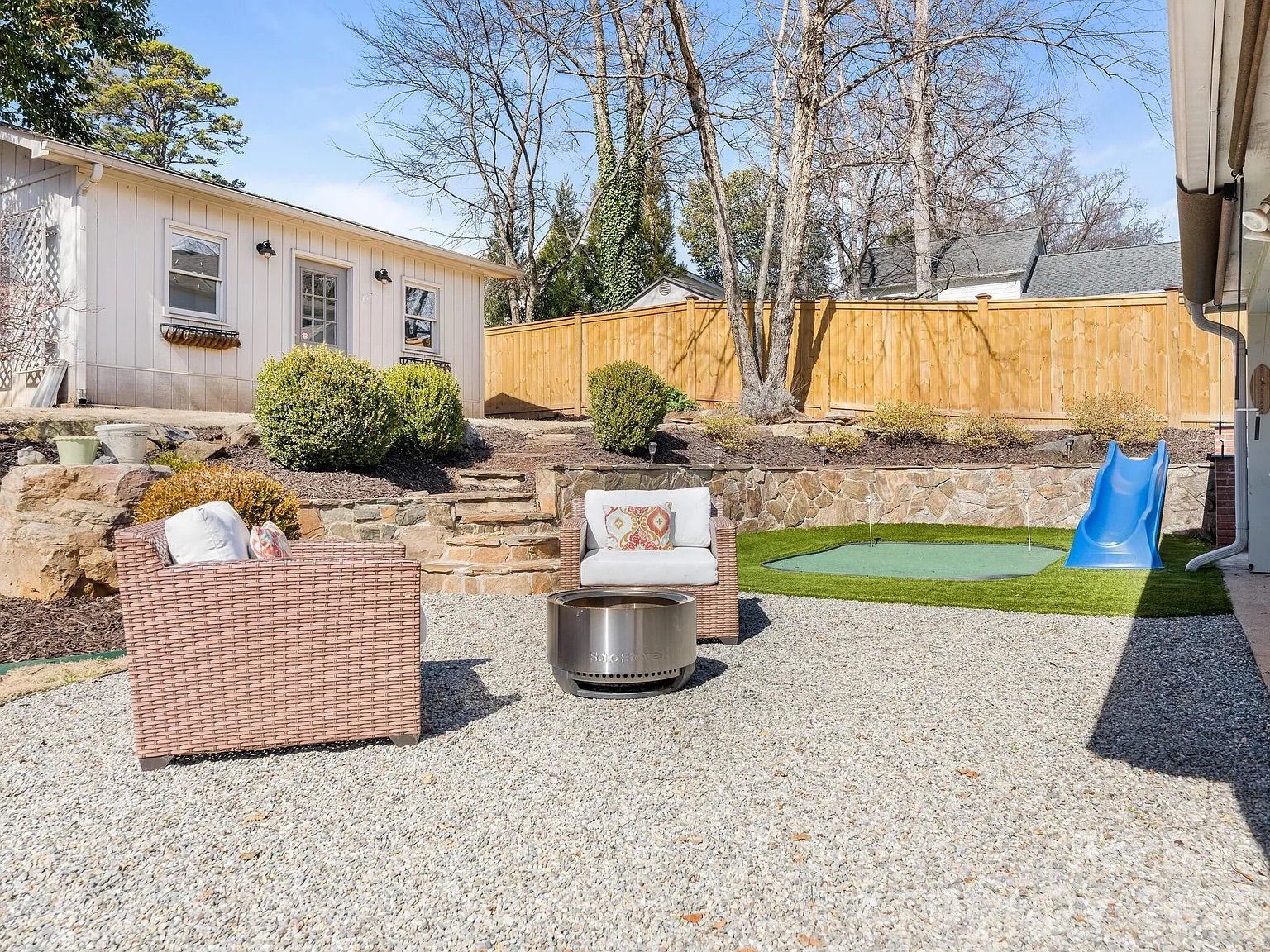 Sunny backyard with two wicker chairs, a round metal fire pit, stone steps, manicured bushes, a wooden fence, a green putting green, and a blue slide.