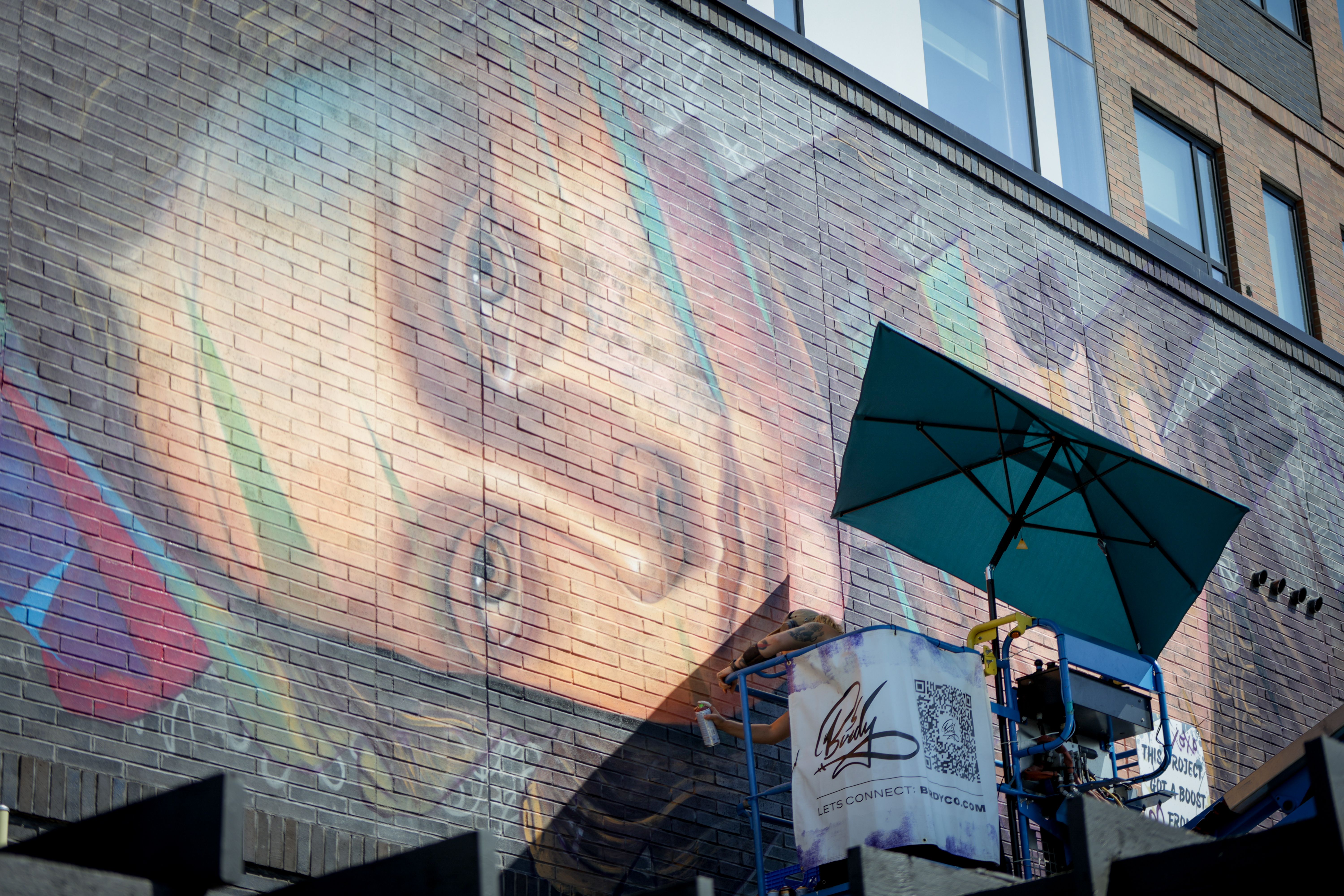 Brick wall mural of Mona Lisa and blue-green highlights; windows above, teal canopy in foreground, and scaffolding with banners.