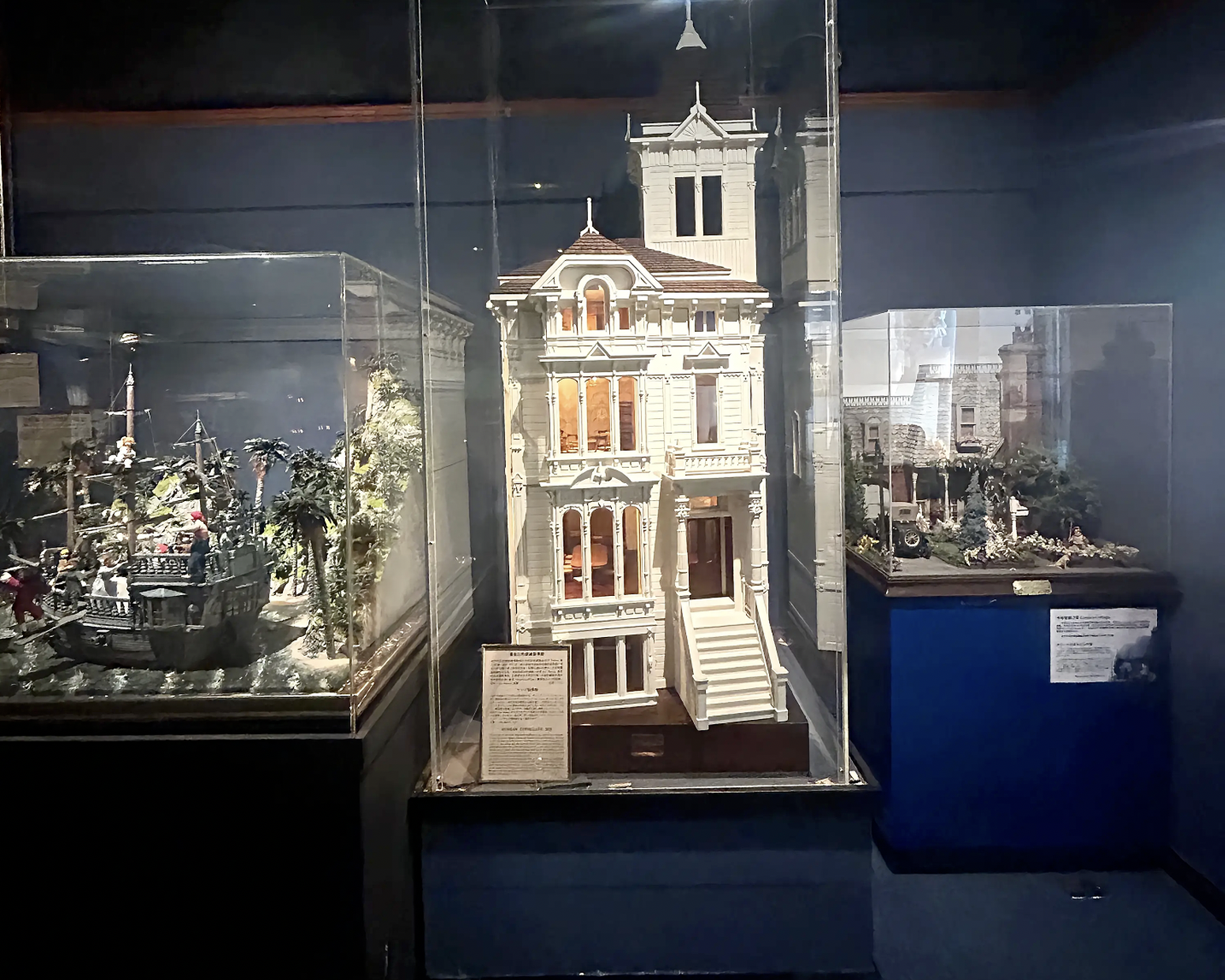 Display of a tall white Victorian-style model house with a stepped facade and small tower behind, set in glass cases with tropical diorama scenes on the left and a ruined building on the right.