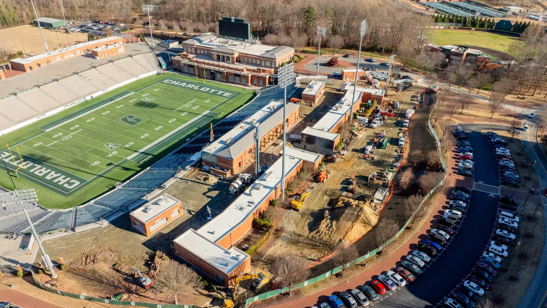 Aerial view of Charlotte football stadium with green field and white lettering, surrounded by brick buildings and ongoing construction, parking lot with parked cars, and trees.