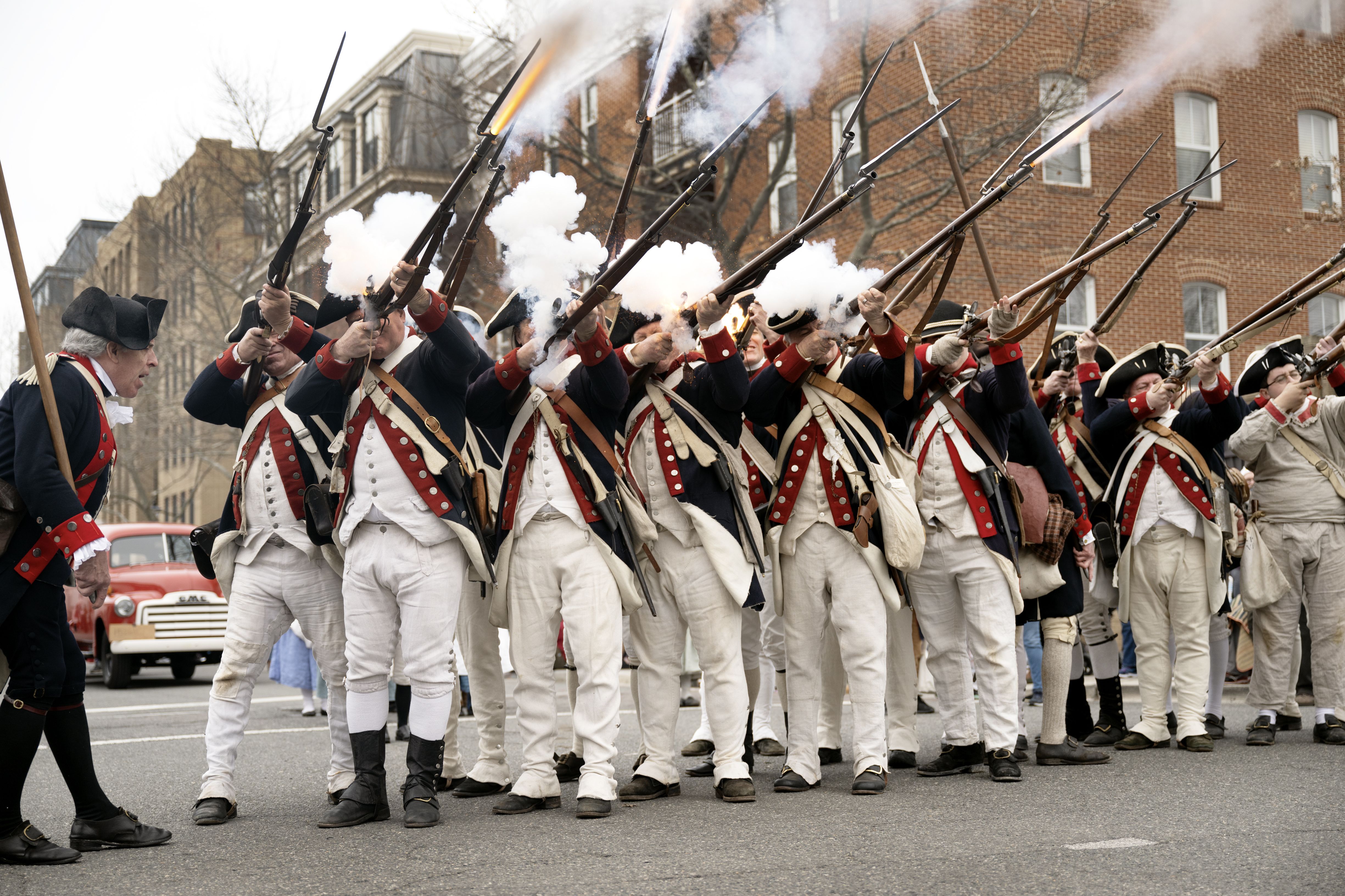 Group of men in historical military uniforms with black hats, red and blue coats, firing muskets with smoke and flames visible, standing on a city street during a reenactment.