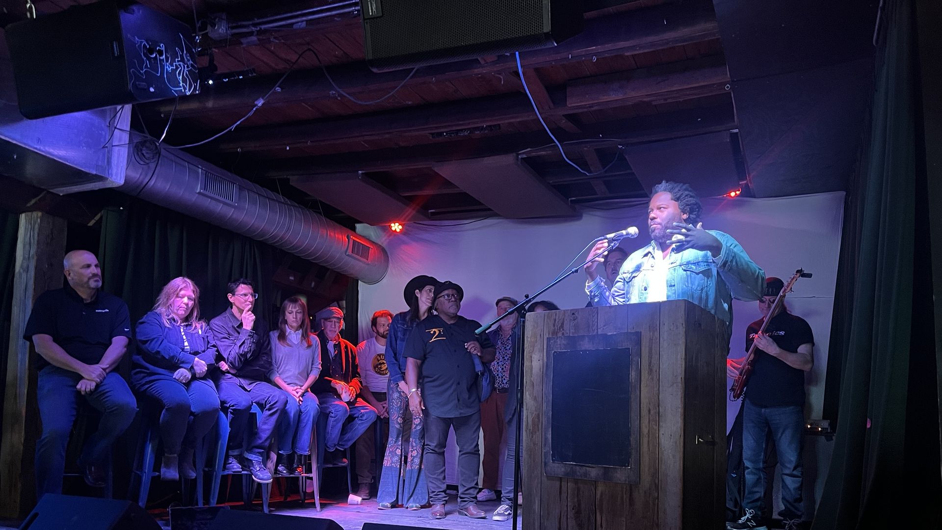 Man with a beard speaks into a microphone at a podium on a small stage; wearing a denim jacket. A row of audience members sits to the left in a dim, purple-lit venue with exposed ceiling lines.
