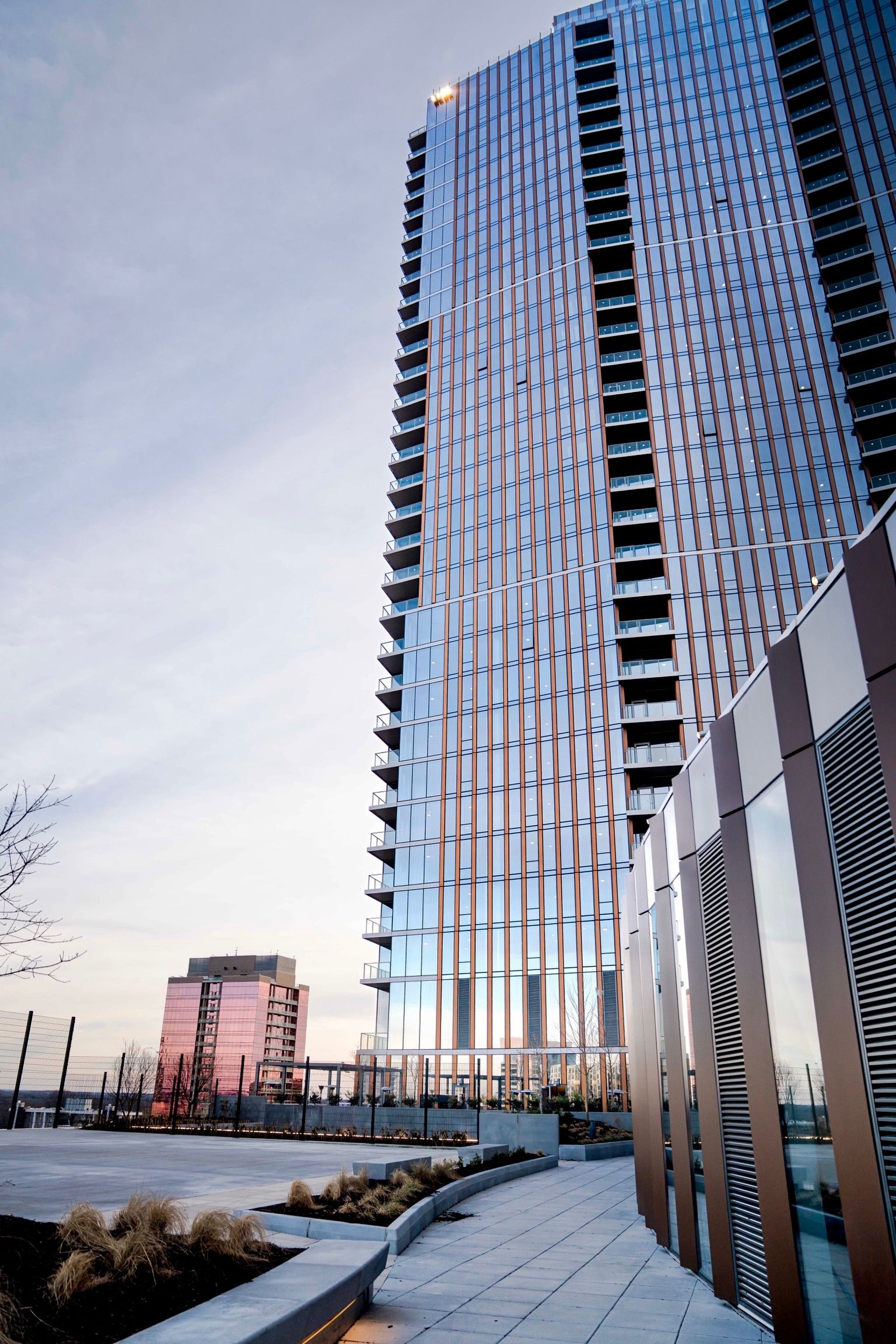 Tall modern glass building reflecting the sky, with a smaller building in the background and paved walkway with plants in the foreground during dusk with soft light.