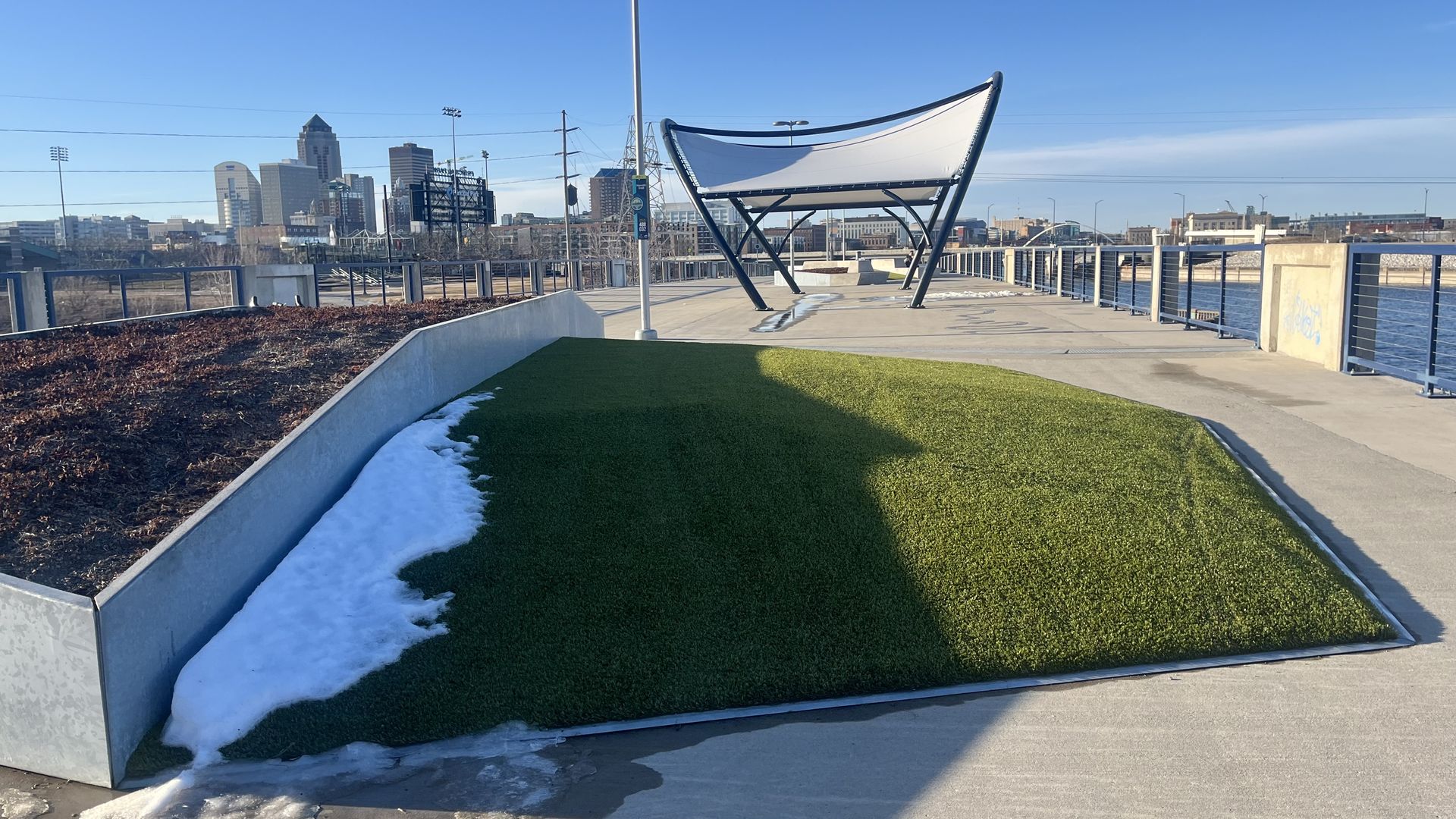 Sunny waterfront promenade with a curved white canopy over a concrete path, a green artificial turf mound, remaining snow along the edge, metal railing, and a distant city skyline under a clear blue sky.