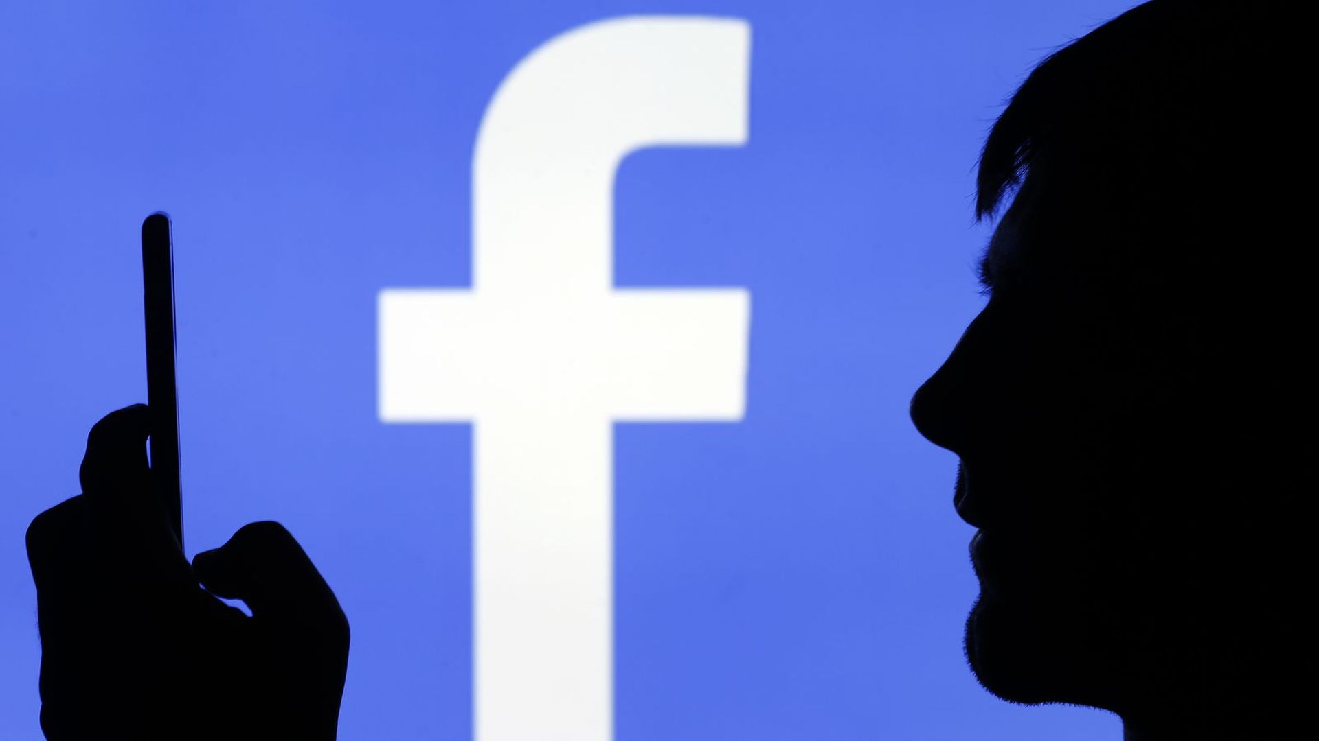A photo illustration with the silhouette of a man on a smartphone in front of a Facebook logo.