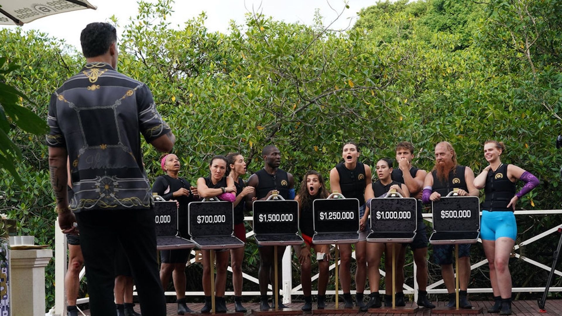 six briefcases are open showing dollar amounts inside, multiple people stand behind the row of briefcases looking shocked