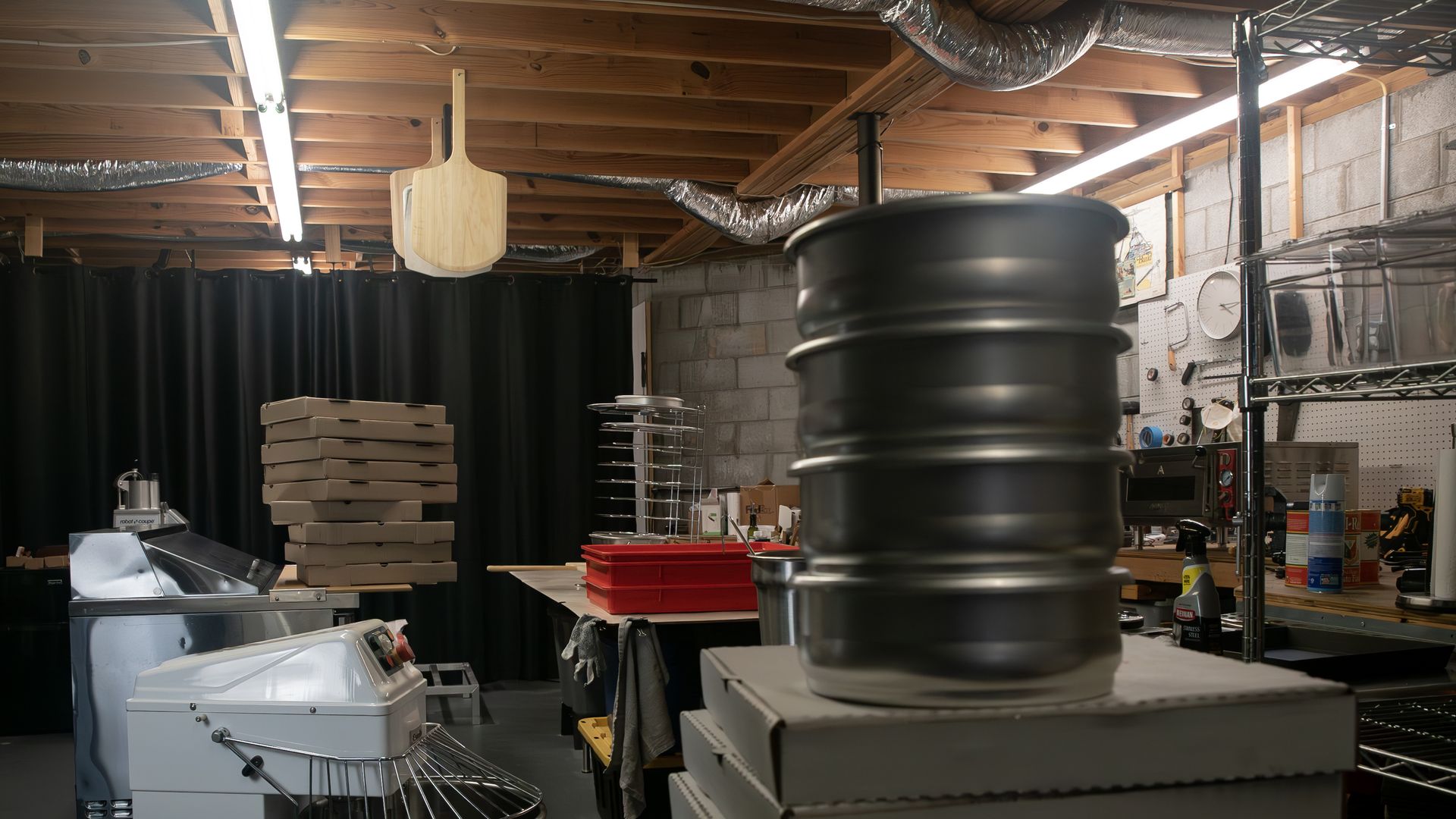 Industrial workshop with exposed wooden ceiling and ductwork, fluorescent lights, a wooden pizza peel hanging, stacks of pizza boxes, metal drums, and pegboard wall with a clock.