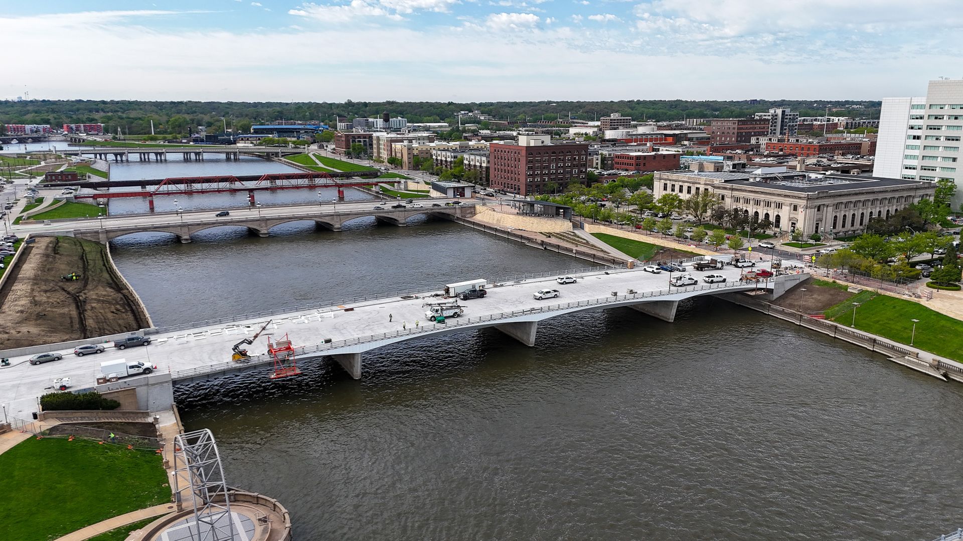 Panoramic aerial view of a river city: multiple bridges cross a wide river, red steel truss bridge and stone arches, brick buildings, green parks along the banks, and a large white building on the right.