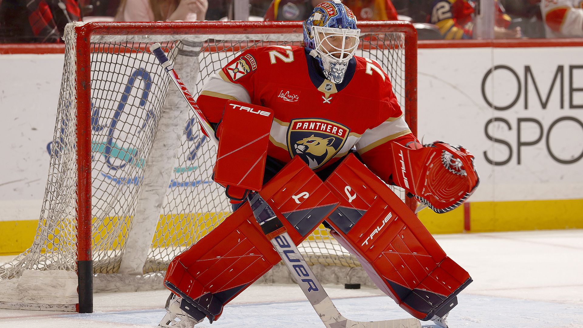 A hockey goalie clad in a mostly red uniform with a Florida Panthers logo stands ready in goal, holding his stick at ice level. 