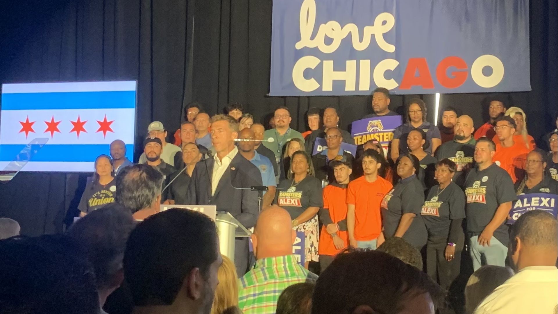 A crowd of diverse people on a stage, a speaker at a podium. Behind them, a blue banner reads "Love Chicago" and a screen shows the Chicago flag with red stars.